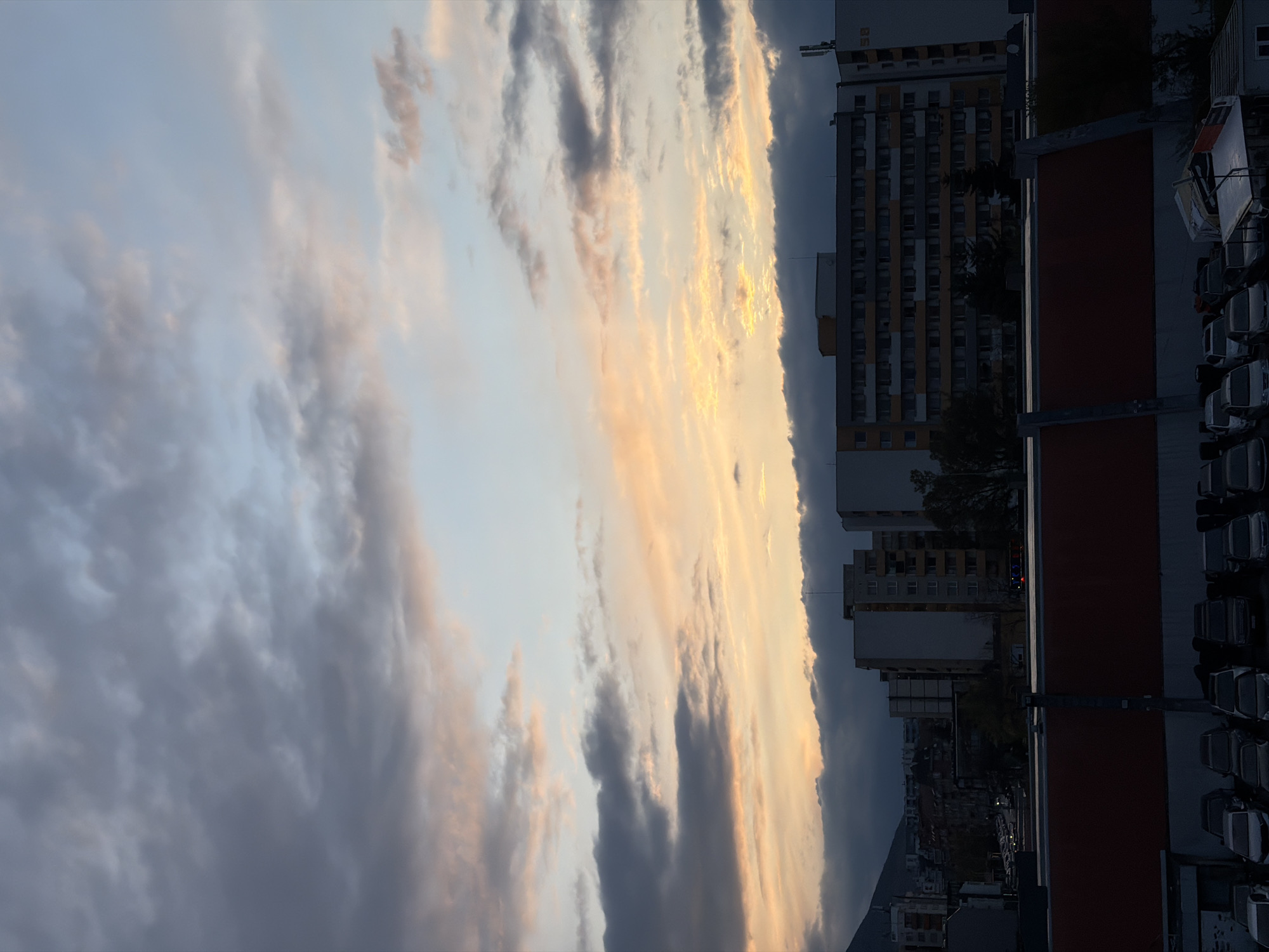 The image depicts a cityscape under a dramatic cloudy sky, likely at sunset or sunrise, with Sofia, Bulgaria as the location context.
The upper two-thirds of the frame are dominated by the sky and clouds. On the left, the clouds are dark grey and blue, gradually transitioning towards the right to vibrant golden, orange, and peach hues, illuminated by the low sun. Some clouds appear wispy, while others are dense and volumetric.
The lower third of the image shows urban elements. On the right, several tall, multi-story apartment buildings are visible, some partially silhouetted against the bright sky. These buildings feature numerous windows and some incorporate yellow and grey facades. In the foreground on the right, a dark red or brown flat rooftop structure stretches across, and behind it, a parking area with multiple light-colored cars (white, silver, grey) is visible in rows. Dark trees and vegetation are interspersed among the buildings. In the far distance on the left, faint outlines of more buildings and a dark hill or mountain range can be seen beneath heavier cloud cover.
The time of day is characterized by the warm, glowing light on the clouds, strongly indicating either a sunrise or sunset. The weather is cloudy, but with clear enough atmospheric conditions to allow for a colorful sky. No specific activity or event is taking place; it's a static urban landscape view. On the upper part of one of the taller buildings, faint letters, possibly "BE" or "8E", are discernible. The architectural style is consistent with post-socialist era buildings common in Eastern European cities.