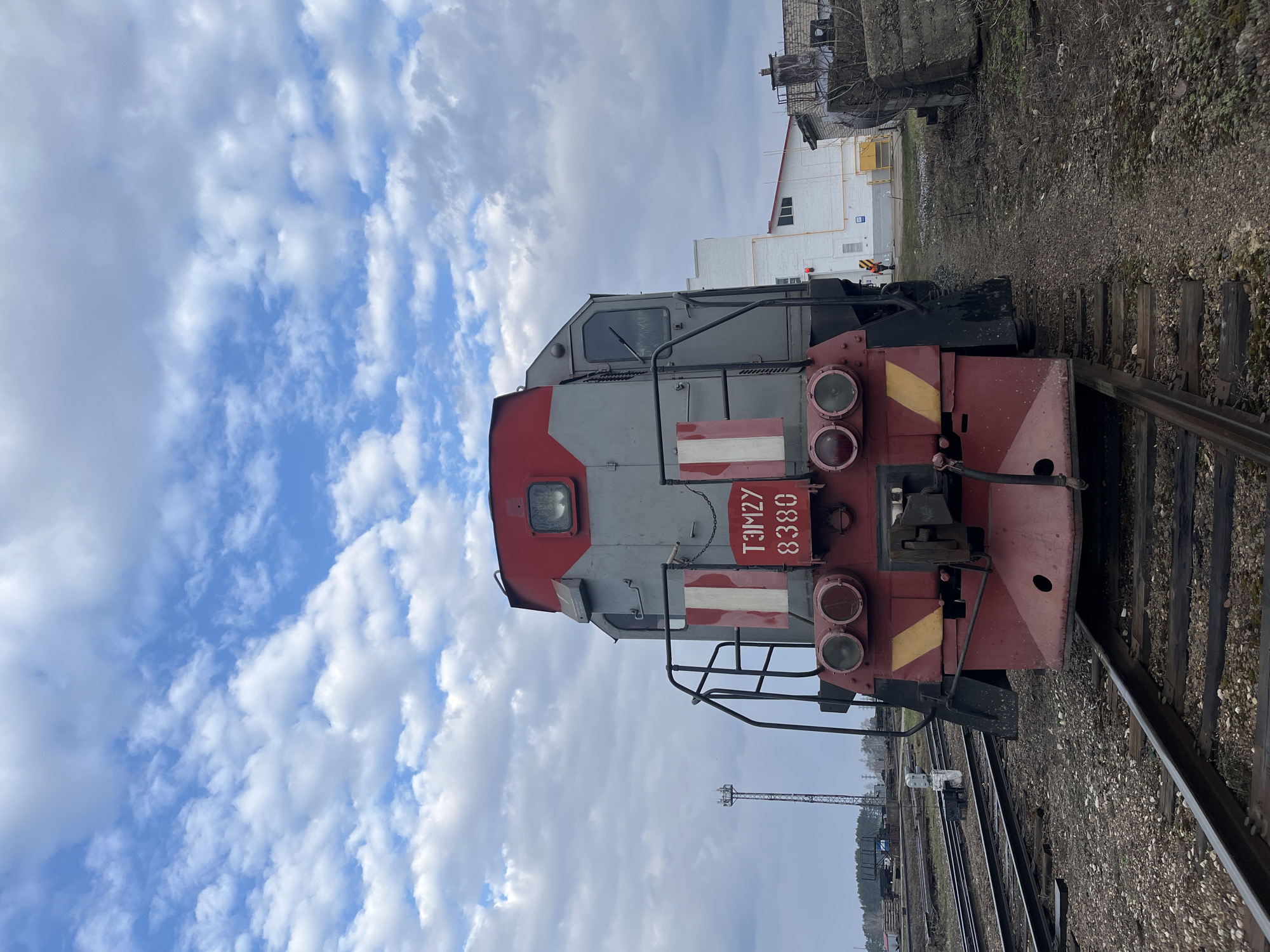 A TGM23 locomotive, specifically TGM23B-8380, is parked on a railway track in what appears to be a railway yard. The sky overhead is cloudy with patches of blue. A white building and other industrial structures are visible in the background. Given the locomotive model information (TGM23B-8380), the location is very likely the yard of the Uralorgsintez plant in Chaikovsky, Perm Krai, Russia.