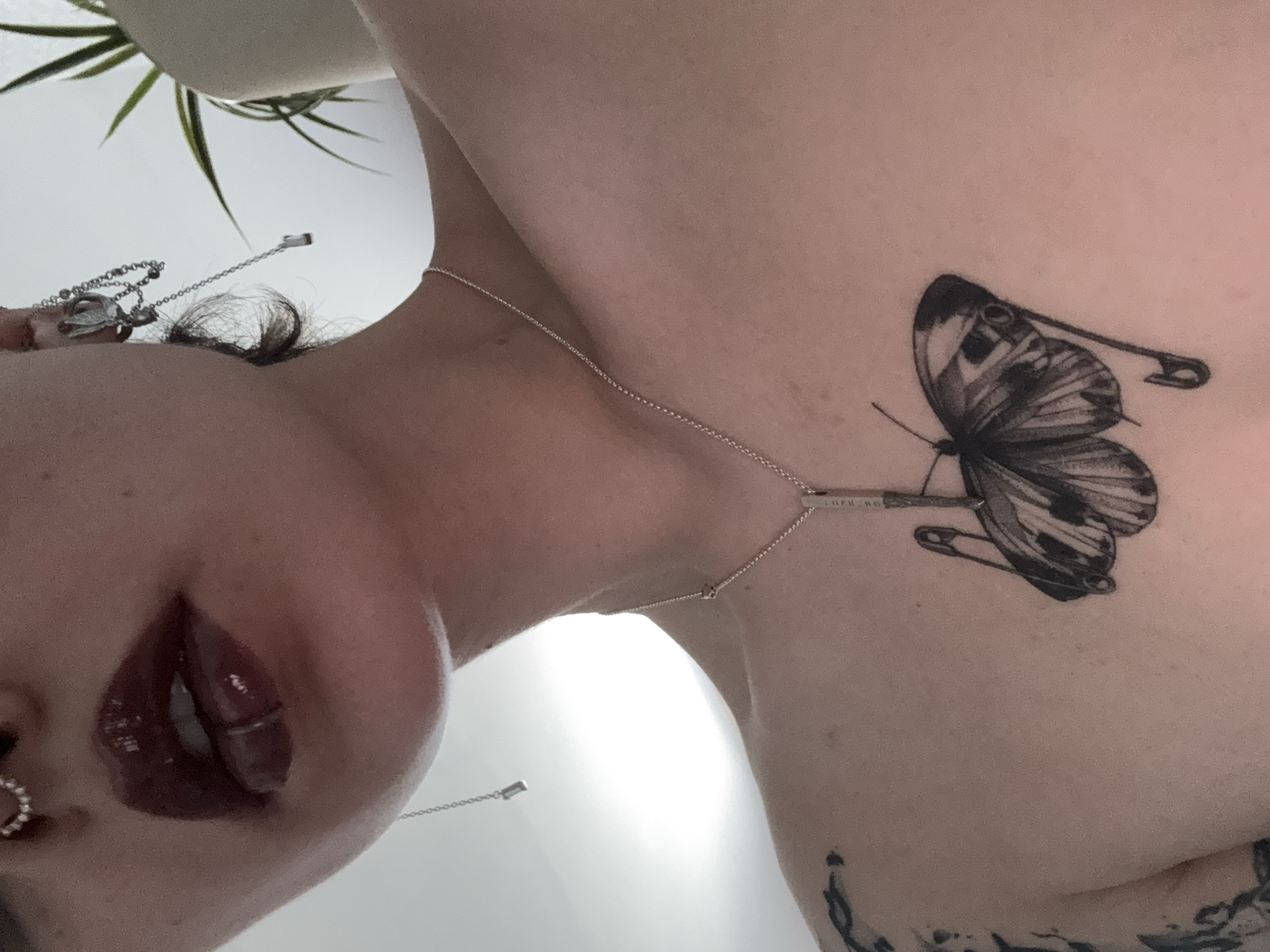 A close-up, low-angle shot of a person tilted at an angle, revealing their face and upper torso. The person has dark, glossy lips, and a nose ring is visible. A delicate silver chain necklace adorns their neck, and another necklace hangs down. A tattoo of a butterfly impaled with a safety pin is visible on their upper chest. The background is a plain white surface with a plant peeking in from the top left corner. The location is not identifiable as being in St. Petersburg, Russia, based solely on the image.