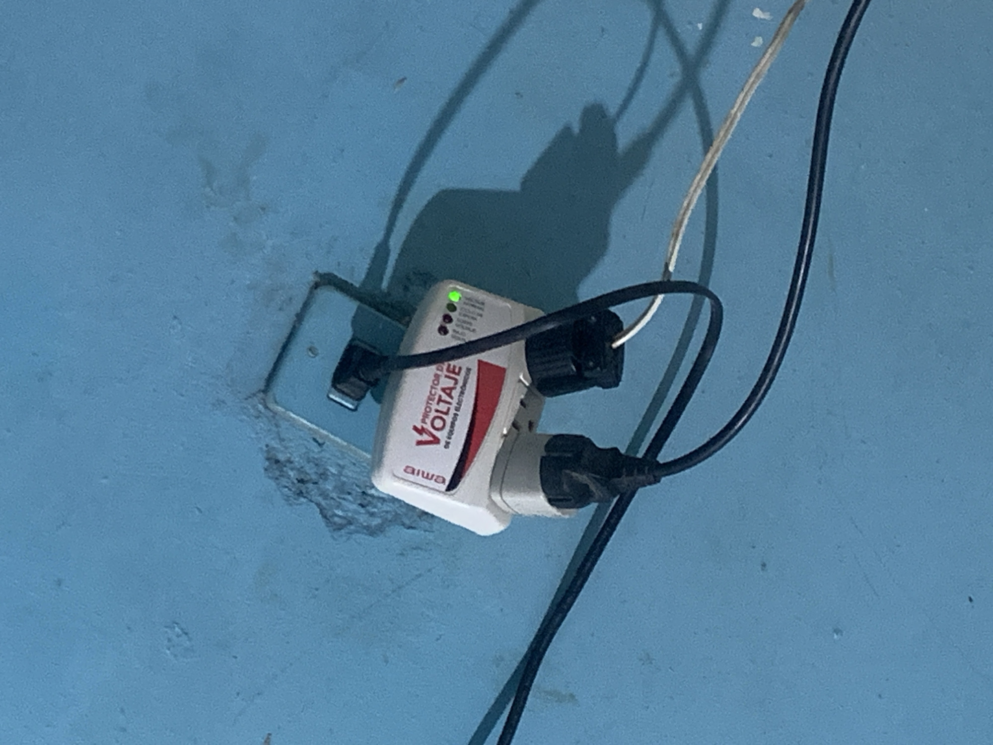 The image captures a white electrical surge protector plugged into a light blue wall outlet on a blue-painted wall in Santa Ana, Venezuela. The surge protector is branded "aiwa" and features the text "PROTECTOR DE VOLTAJE DE EQUIPOS ELECTRONICOS" (Voltage Protector for Electronic Equipment). A green LED light is illuminated on the device, with several unlit red circular indicators visible below it. Two black electrical plugs are inserted into the surge protector, with multiple black and one white electrical cables extending from these connections. The area around the wall outlet shows signs of wear and chipping. Distinct shadows of the cables and the surge protector are cast onto the blue wall, suggesting the presence of a light source. The scene depicts a functional electrical setup for protecting electronic devices.