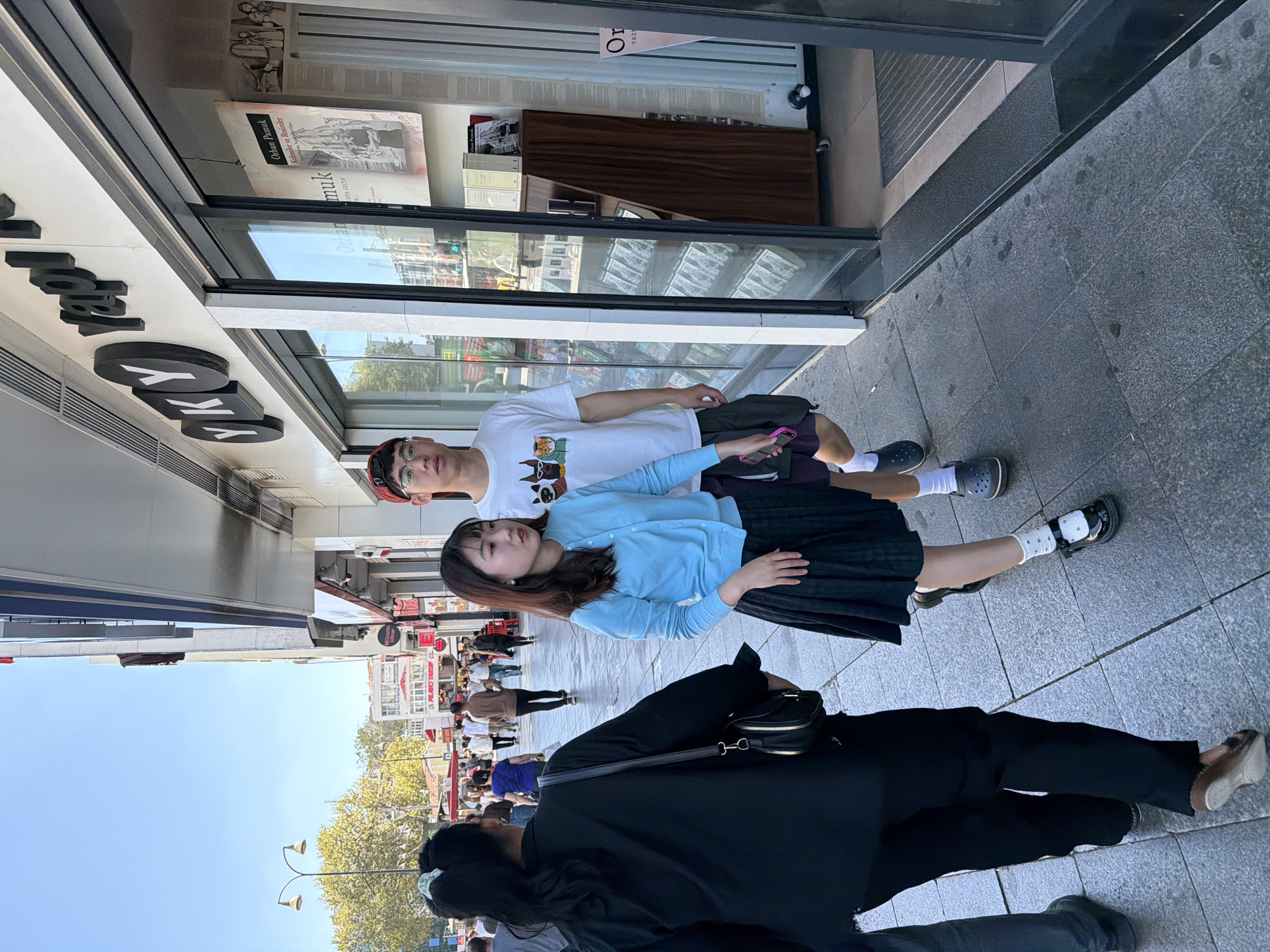 The image depicts a street scene in Kadıköy, Turkey, with a focus on a group of individuals walking on a sidewalk in front of a store. The store's facade features a large window display and signage with the letters "YKY Yapı." Several people are visible in the foreground and background, suggesting a busy urban environment.
In the foreground, a young woman in a light blue cardigan and a pleated black skirt is walking towards the viewer, accompanied by a young man wearing a white t-shirt with a graphic print and glasses. To the left of them, a woman with her back to the camera is wearing a black jacket and carrying a black purse. Their path leads down a sidewalk paved with grey tiles.
The store behind them has a large glass entrance and display windows showcasing books and other merchandise. The exterior signage indicates it is likely a bookstore or similar establishment. The street appears to be a commercial area, with other shops and buildings visible in the background. The lighting suggests it is daytime, with clear skies. The overall impression is that of an ordinary day in a bustling Turkish city.