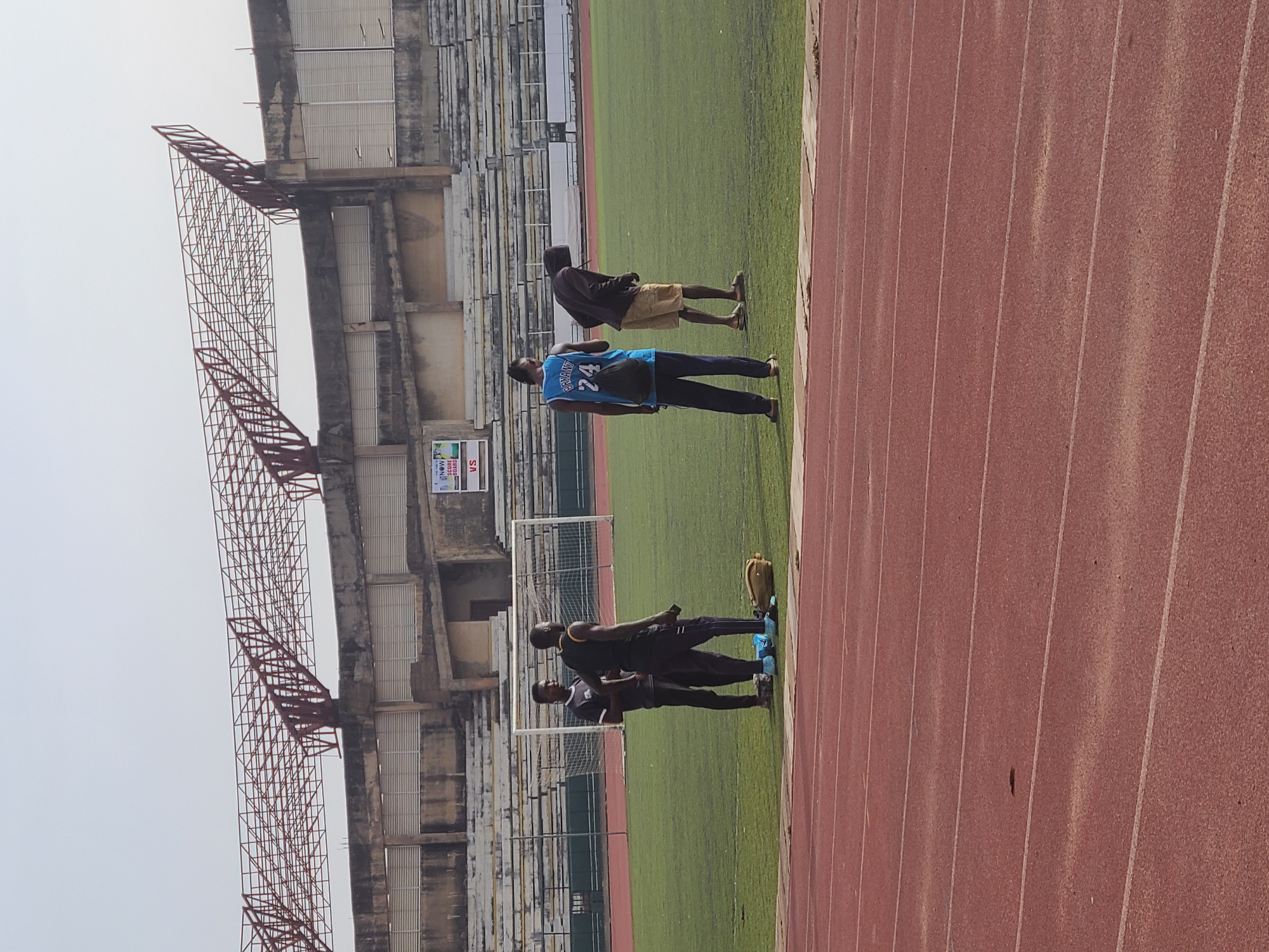 The image shows a soccer field, presumably the New Stadium in Nigeria, with several people standing along the edge of the running track. A man in a blue jersey with the number 24 is prominent. There is a partially constructed stadium in the background and goal posts on the field. A tan backpack is on the green turf.