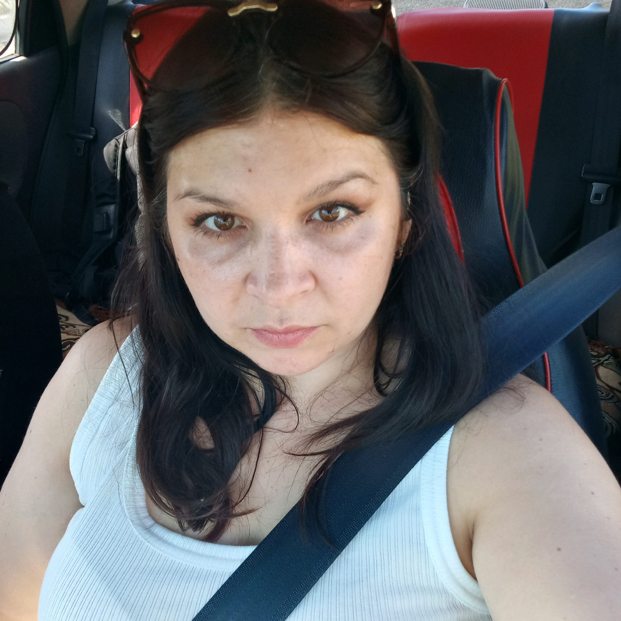 In a frontal selfie, a woman with dark hair and brown eyes sits in the passenger seat of a car, wearing a white tank top and sunglasses perched on her head. The interior of the car is visible, featuring red and black upholstery. A blue seatbelt is fastened across her chest. The background suggests an outdoor setting, possibly a street or residential area. The image does not contain any information that places it in Klyntsi, Ukraine.