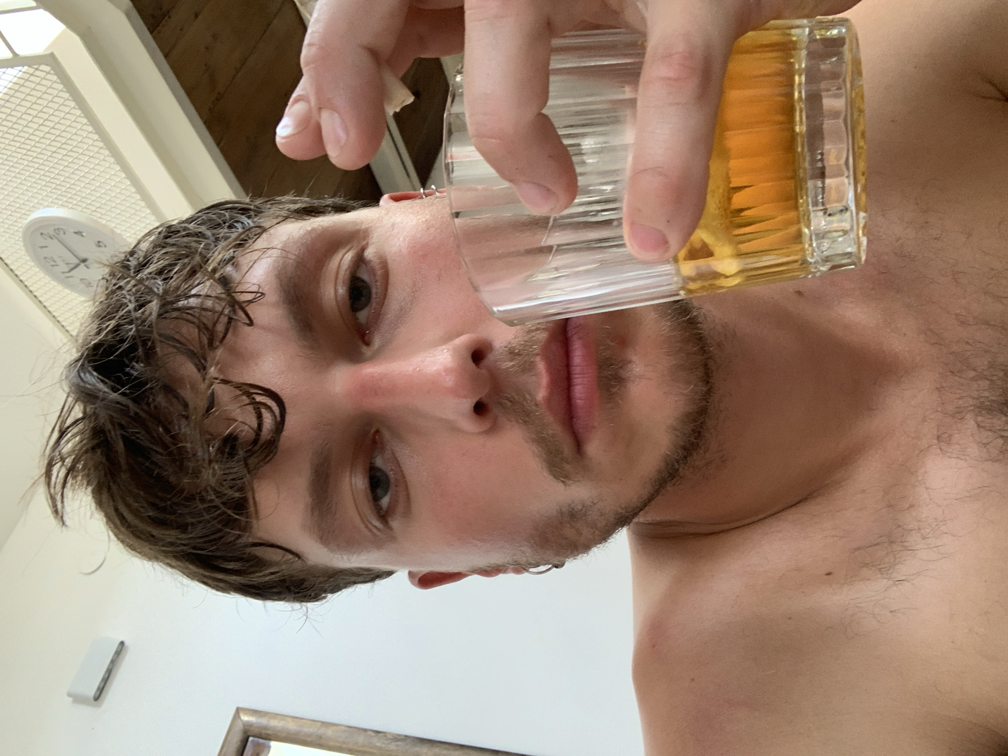 An overhead shot captures a young man, seemingly in a relaxed indoor setting, holding a clear, ribbed glass filled with a golden-yellow liquid, possibly a beverage, near his mouth. He is shirtless, revealing a fair complexion and some chest hair, along with stubble on his chin and a mustache. His dark, wet, curly hair is styled somewhat casually, and he has a silver-colored hoop earring visible in his left ear. He looks directly into the camera with a neutral, possibly contemplative, expression.
In the background, a white analog clock is mounted on a light-colored wall, showing the time to be approximately 2:40 or 2:50. Above the clock, a wooden ceiling or beam is visible. To the bottom of the frame, part of a gold-framed mirror can be seen. Another white rectangular object is affixed to the wall below the clock. The lighting suggests daylight. There are no specific visual cues within the image to indicate a precise geographical location. The numbers "12" and "10" are clearly visible on the clock face.