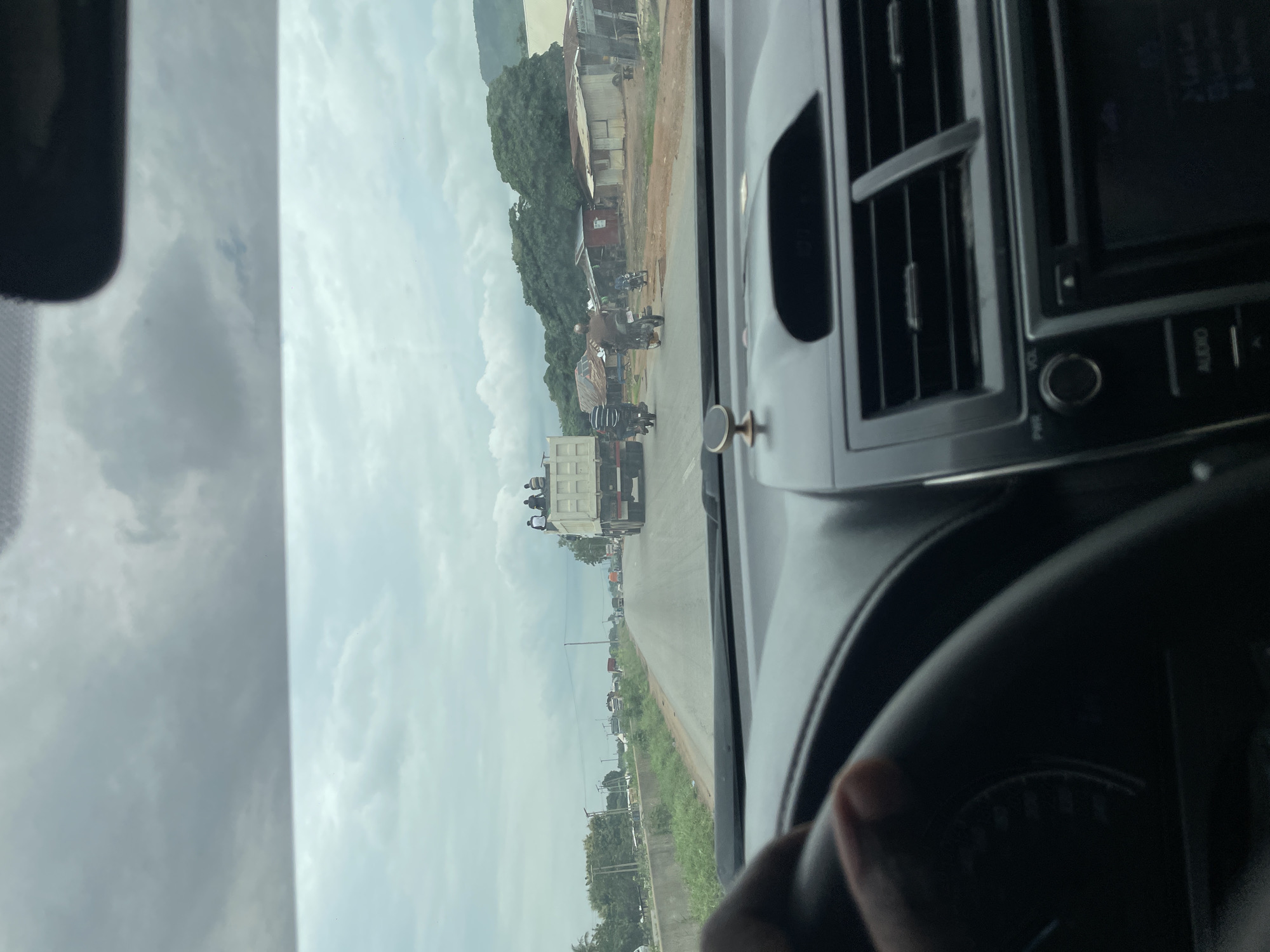 This media file captures a view from the interior of a vehicle, likely a car, as it travels along a road. The scene outside the windshield depicts a rural or semi-urban setting in Kwali, Nigeria.
The primary subjects visible are a series of vehicles on a road, including a large white truck in the foreground loaded with passengers, several motorcycles, and other cars further down the road. There are also buildings and trees lining the road. The sky is cloudy, suggesting overcast weather, and the overall lighting indicates it is daytime. The perspective suggests the vehicle is moving, contributing to a sense of travel.
Notable details include people riding on top of the white truck, which is a significant visual element. The dashboard of the vehicle is partially visible, showing an air conditioning vent and part of the audio system. The road itself appears to be paved, with a grassy verge.
There is no visible text in the image. The activity taking place is vehicular movement along a road, likely a common travel route in the area. The image conveys a sense of everyday life and transportation in the depicted location.