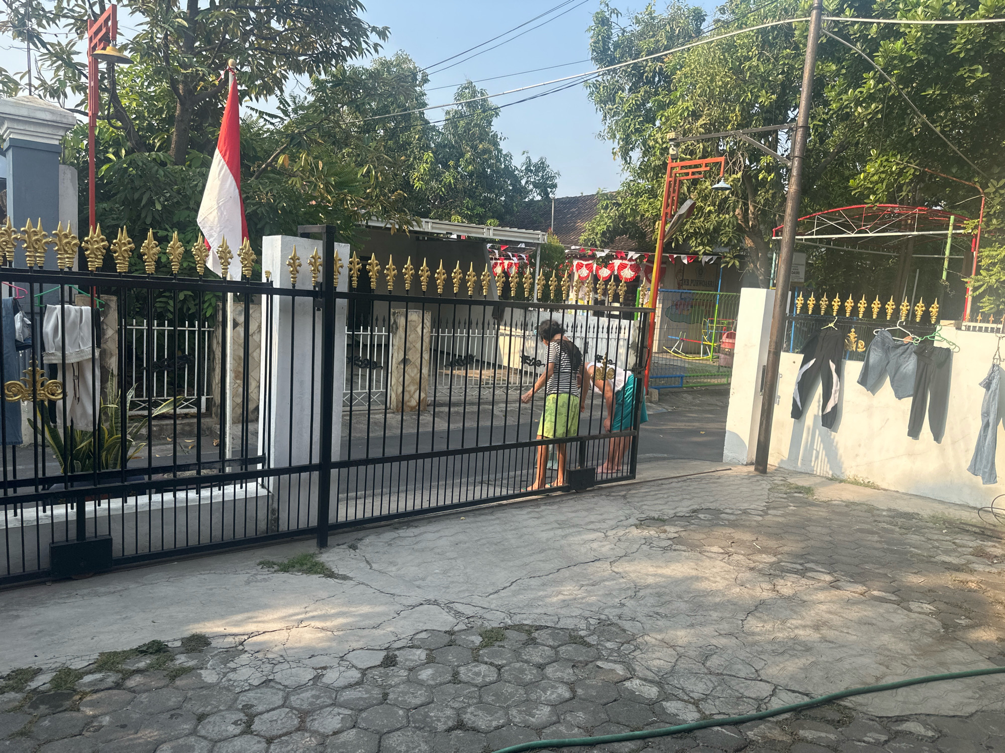 A residential fence in Mojokerto, Indonesia, with a red and white Indonesian flag flying on a pole to the left. The fence is black with gold decorative elements. Inside the fence, a building with a white roof is visible, decorated with red and white pennants. Two individuals are standing near the open gate, with one appearing to be pushing it. The ground outside the gate is paved with concrete and hexagonal stones. Clothes are hanging on a clothesline to the right of the gate. Trees and power lines are visible in the background. The scene is lit by sunlight, casting shadows on the ground.