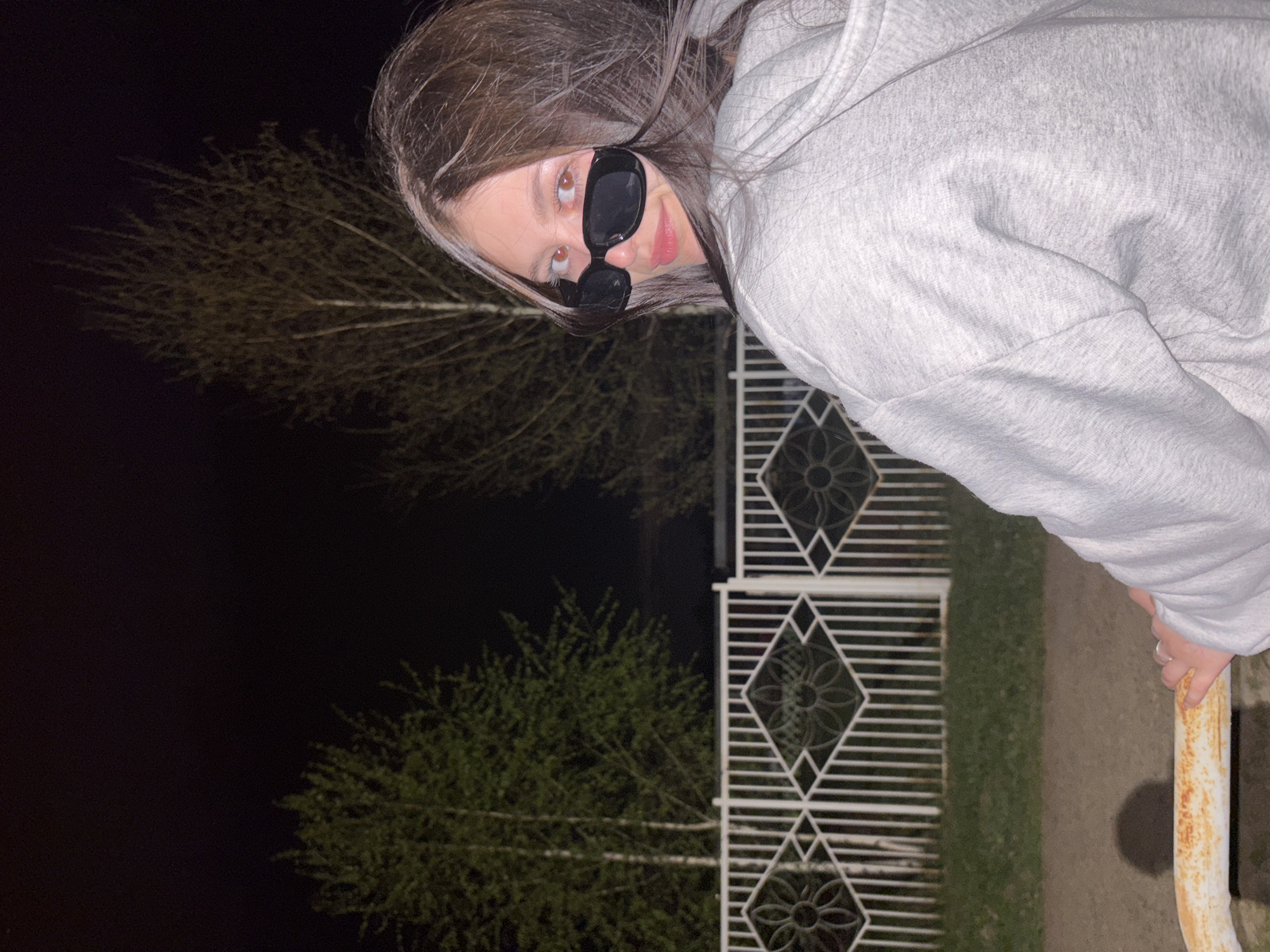 A woman with sunglasses and a gray hoodie is standing in front of a white metal fence with decorative diamond patterns. Behind the fence are two trees, one partially obscured by the darkness. The background is dark, possibly indicating it is night. The woman is looking towards the camera. The location is Фоки, Russia.