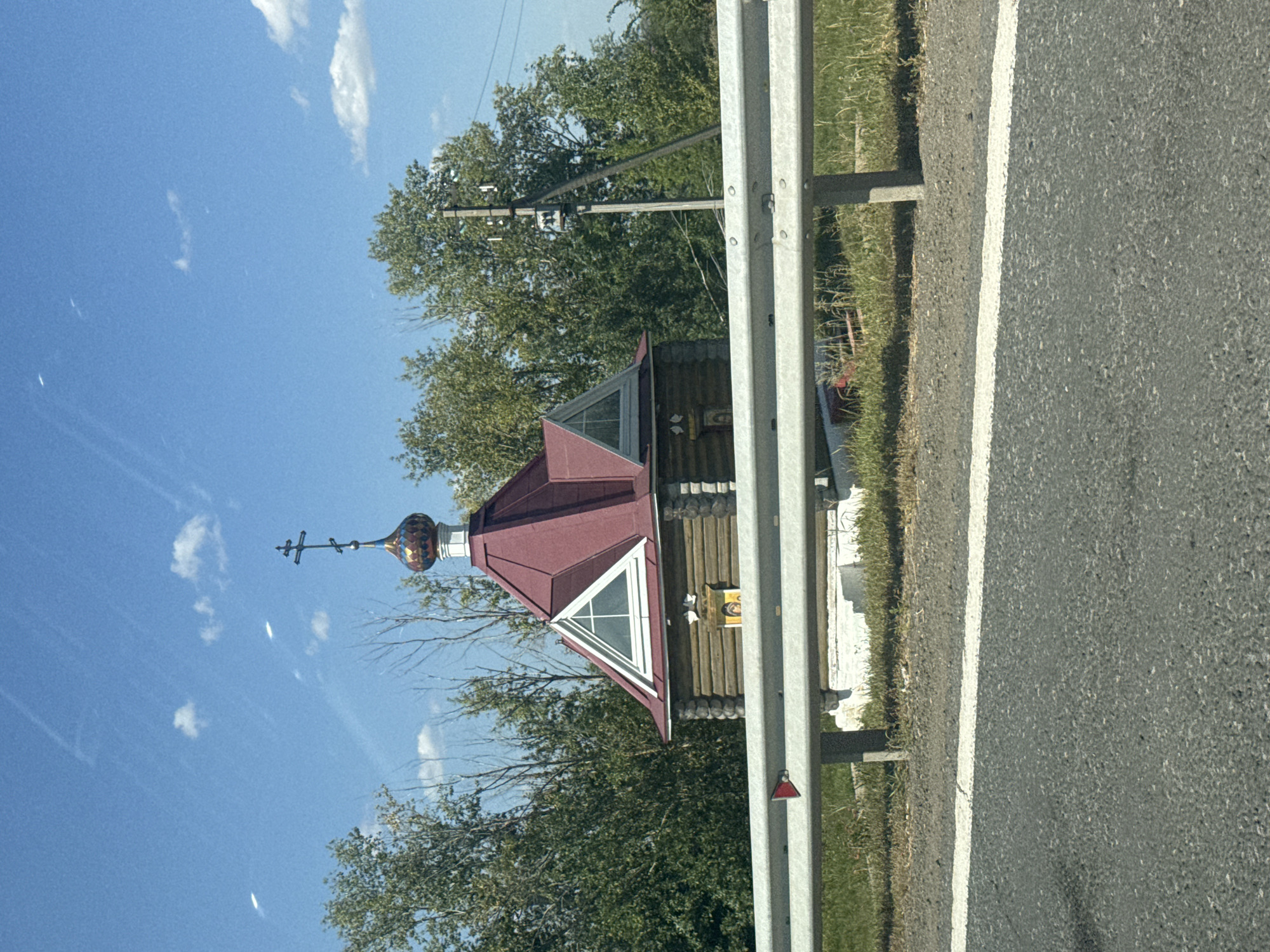 An orthodox chapel with a red roof and a cross stands next to a road in Kholodnye Kliuchi, Russia. The chapel is made of wood with a small icon on the front. A metal guardrail separates the chapel from the road, which has a white stripe marking the lane. Trees surround the chapel, and the sky is blue with some clouds. A utility pole is visible behind the chapel.