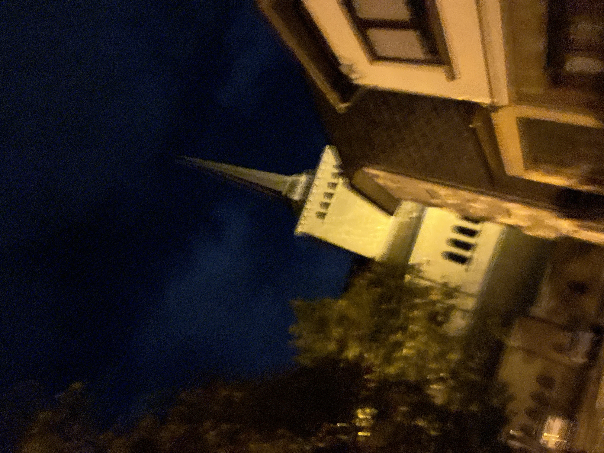 At night, a building with a tall, pointed spire is visible against a dark blue sky. The building is lit by artificial lights, casting a warm yellow glow. In front of the spire building, there is another building with a pitched roof, also illuminated. To the left of the spire building, trees with yellow lights can be seen. The overall scene appears to be in an urban or town setting, possibly Saint-Vith, Belgium, given the architectural style.