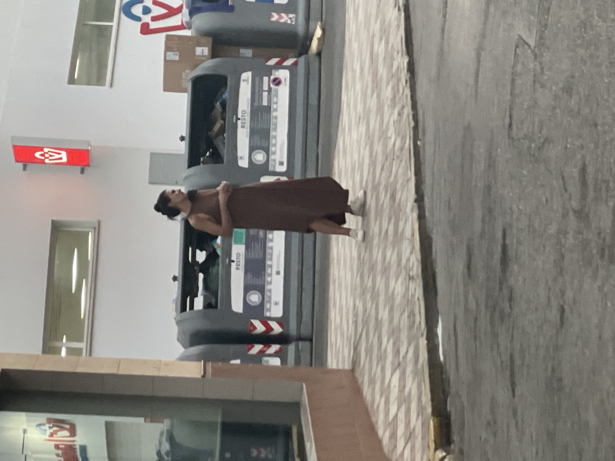 An adult female, with dark hair pulled back into a bun, stands on a tiled sidewalk in front of a row of large, grey recycling bins. She is wearing a long, brown sleeveless dress and white sneakers. The bins are partially full of various items, including what appears to be cardboard boxes and plastic. Above the bins, on a white wall, is a red sign with a white shopping bag logo. To the left of the bins, part of a building facade with a window is visible. The ground in front of the sidewalk is asphalt. The image appears to have been taken in La Línea de la Concepción, Spain, given the context provided by the prompt.