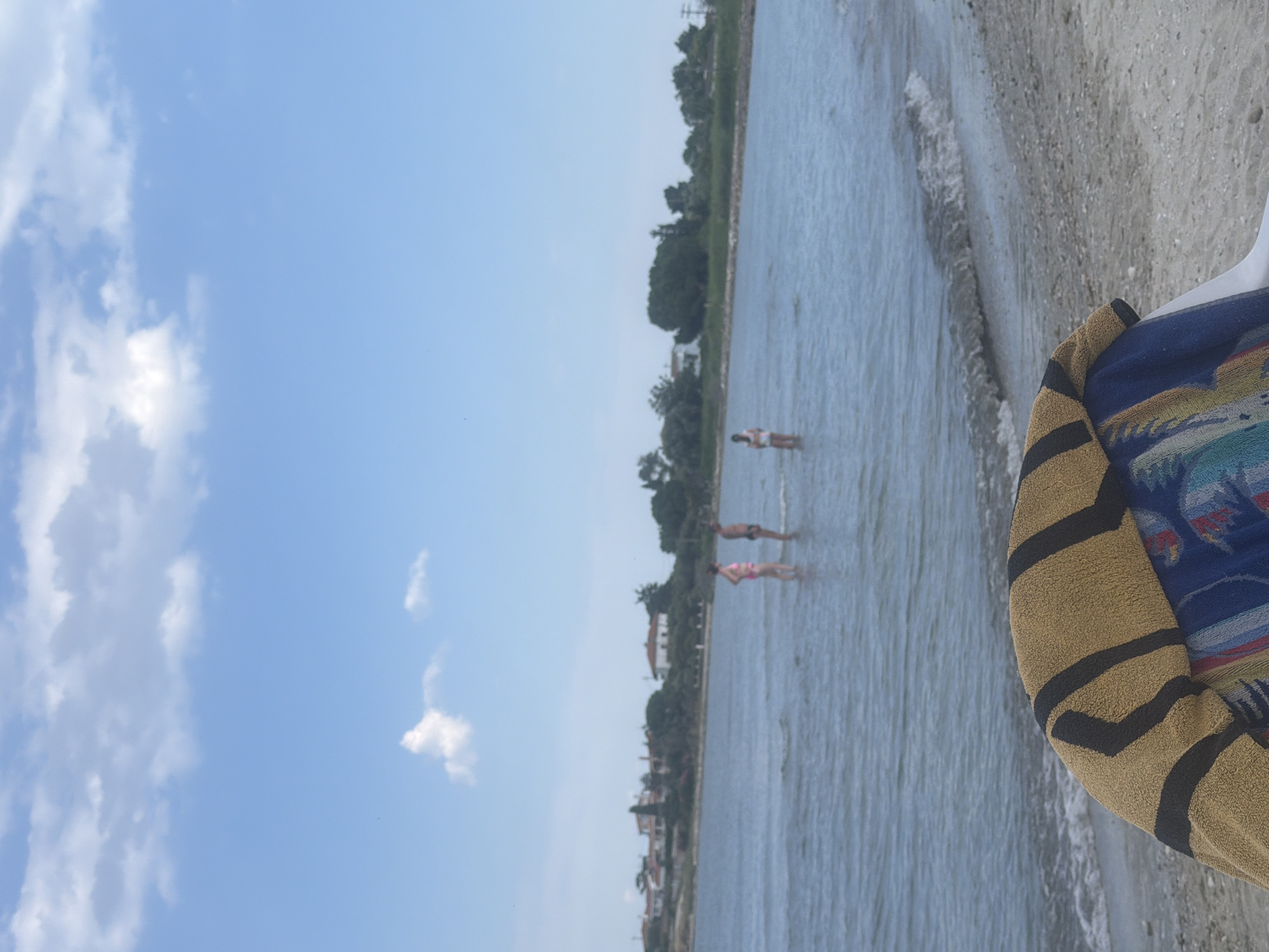 In the city of Mandra, Greece, a wide shot captures a tranquil beach scene. The foreground is dominated by the edge of a beach chair draped with a towel, its textured fabric a mix of tan and black stripes. Beyond, the gentle waves of the sea lap onto the sandy shore. In the shallow water, three individuals are visible. Two people, one in a pink swimsuit and another in dark swim trunks, stand facing each other. Further out, a third person in a white swimsuit stands with their back to the camera. The background reveals a coastline dotted with residential buildings and lush green trees, all under a bright blue sky with scattered white clouds.