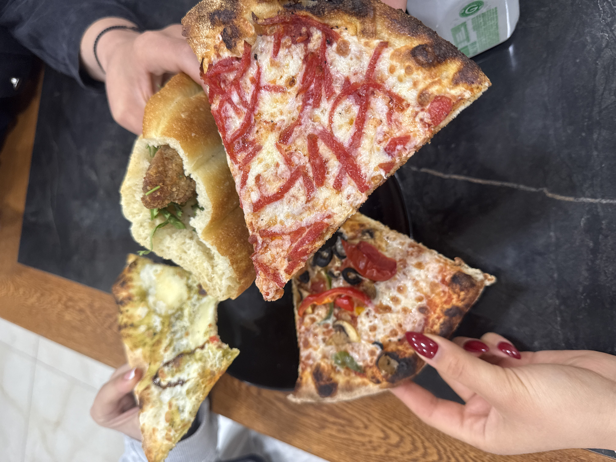The image shows an overhead shot of food items being held by three people. The food includes: a sandwich with a fried patty inside, a slice of pizza with shredded red toppings on cheese, a slice of pizza with various vegetable toppings like olives, peppers, and mushrooms, and a slice of pizza with a greenish topping. The hands holding the pizza slice on the right have red nail polish. The setting appears to be a restaurant or home, with a dark marble-like table and a wooden surface partially visible. The location, Aparan, Armenia, is not discernible from the image.