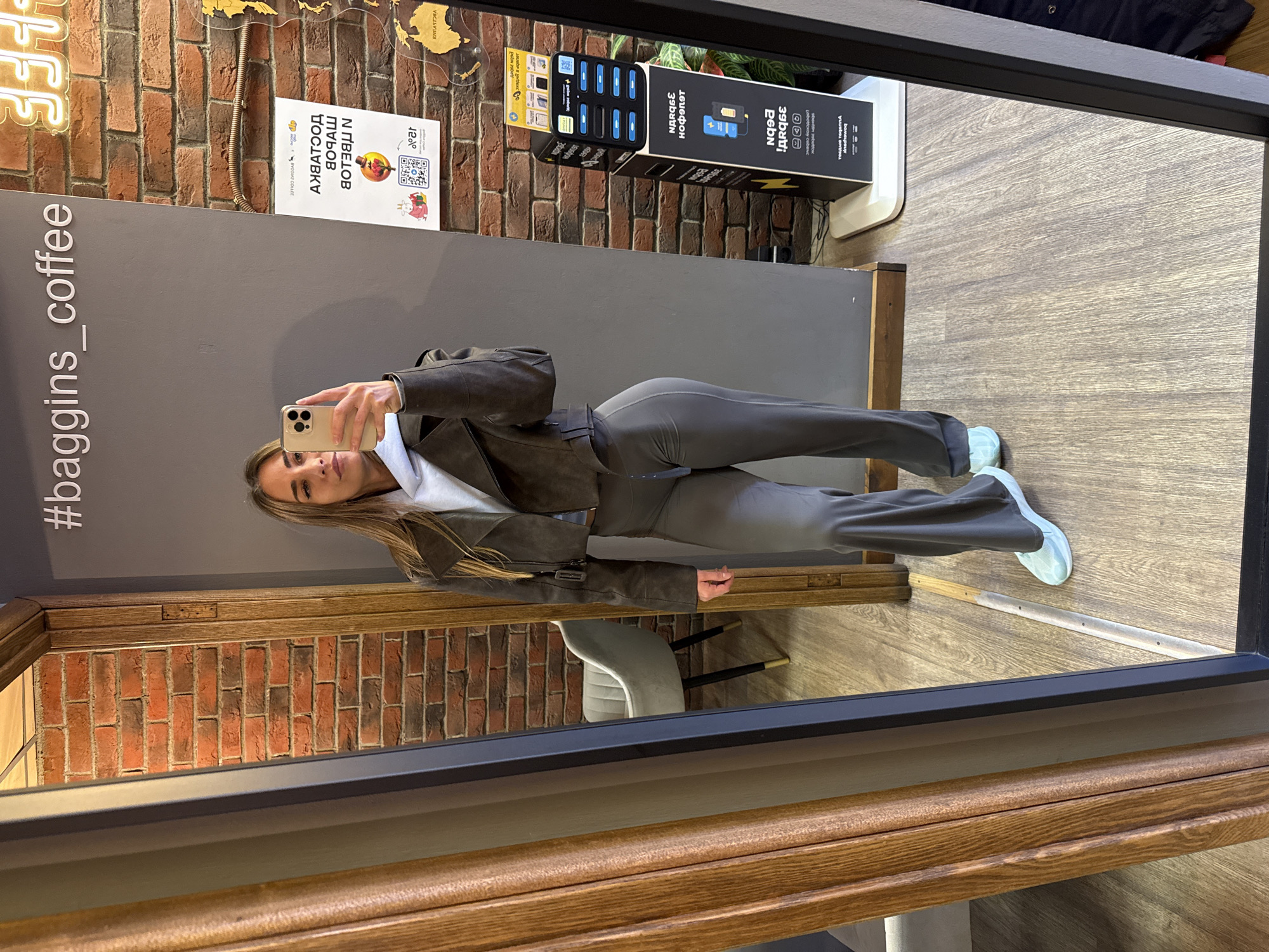 A young Caucasian woman is taking a selfie in a mirror inside a business in Saint Petersburg, Russia. She is wearing a grey athletic outfit and holding a phone. The wall next to her is grey with the hashtag "#baggins_coffee" printed vertically. Behind the wall is a brick wall with neon lighting. A charging station with USB ports is also visible on the brick wall. The floor is made of light wood, and there is a chair reflected in the mirror.