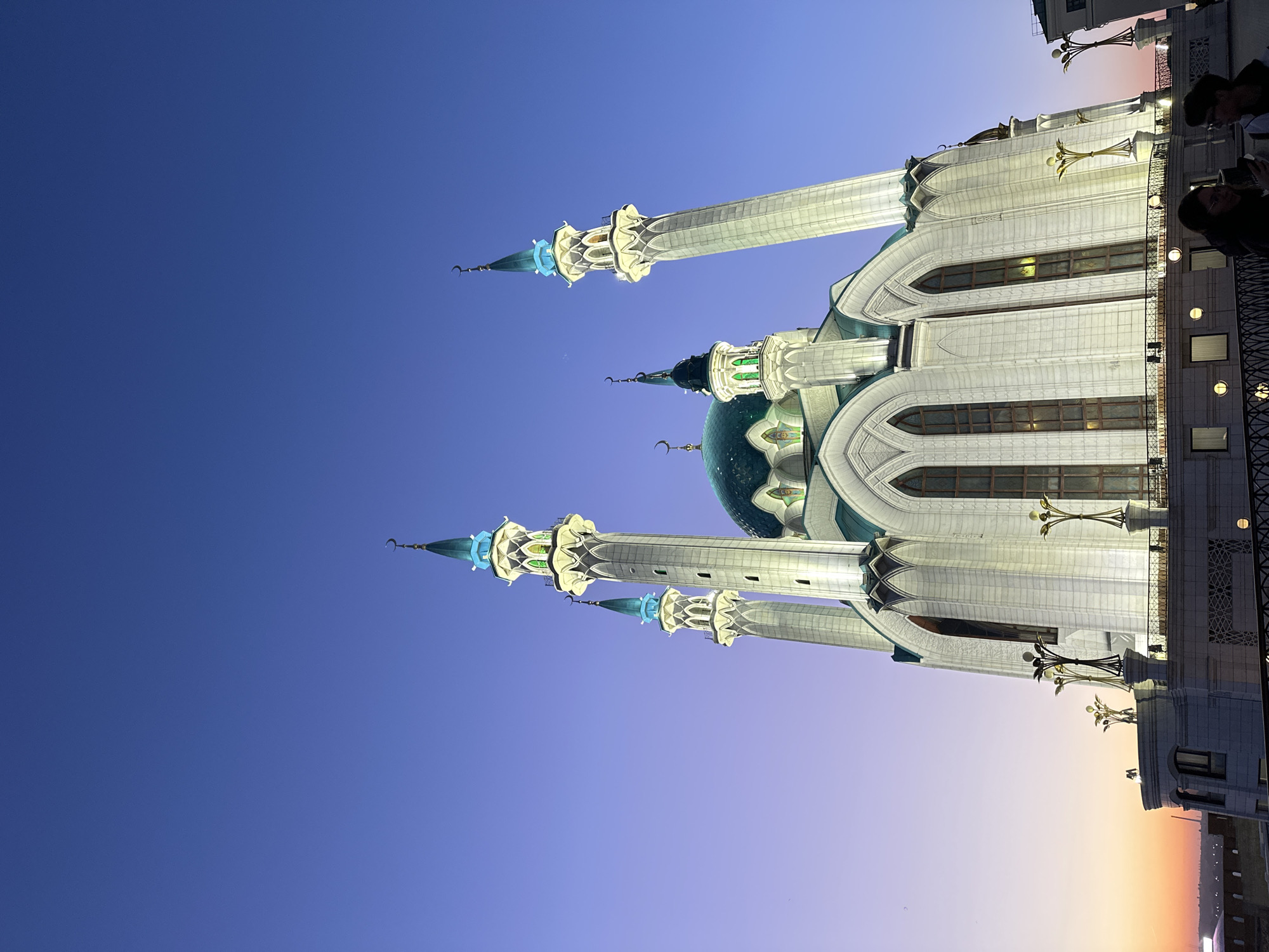 The image shows the Kul Sharif Mosque in Kazan, Russia, taken at dusk. The mosque features white walls, a teal dome, and several tall, slender minarets. The sky transitions from a deep blue at the top to a lighter gradient towards the horizon. A building appears on the right side, with people partially visible on a balcony or rooftop. The mosque is illuminated, with lights highlighting its architectural details.