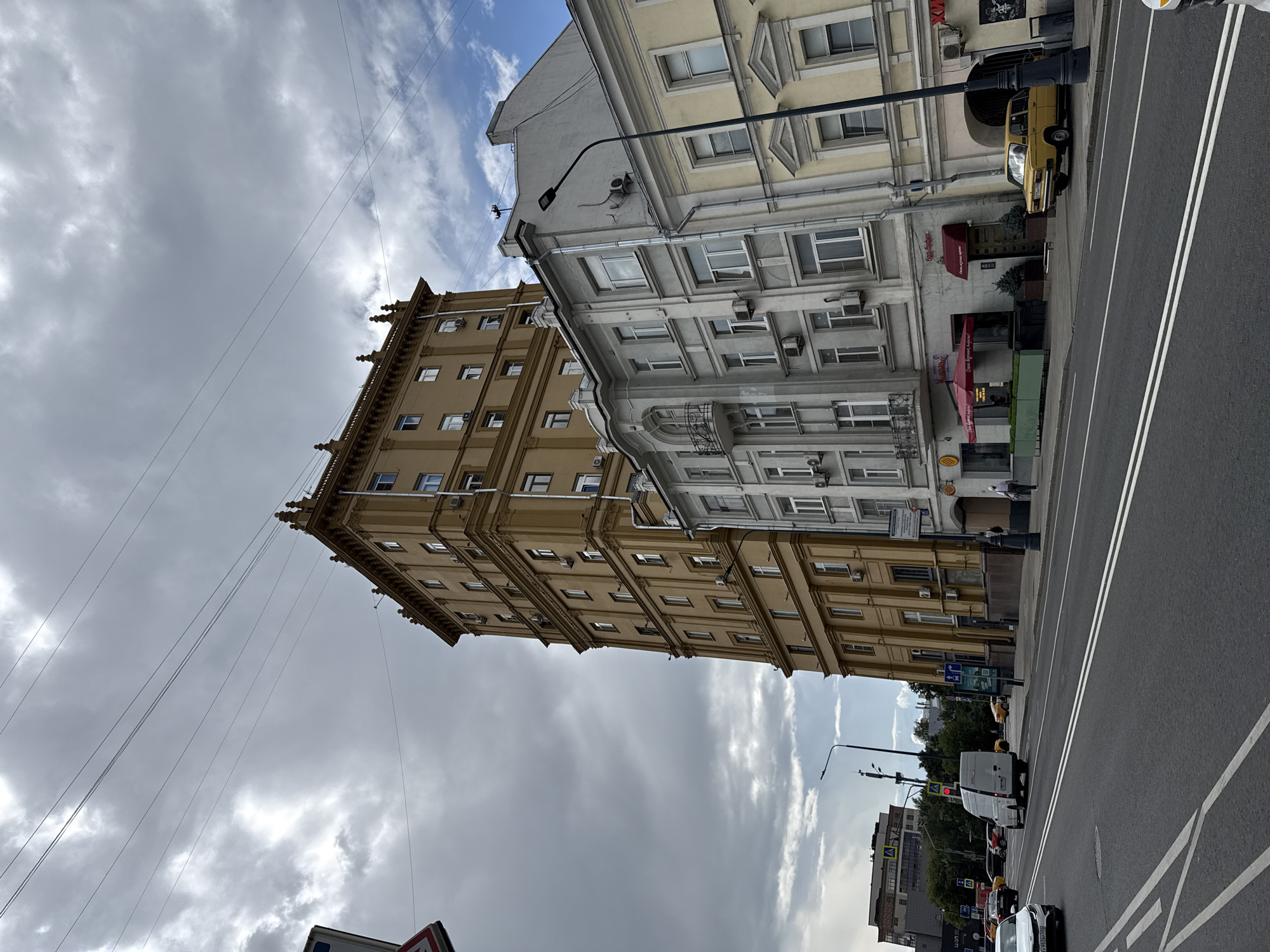 The image captures a street scene in Moscow, Russia, under a cloudy sky. In the foreground, a wide asphalt road with white lane markings runs horizontally across the frame. A white van and several cars are visible on the road. On the right side of the road, a row of buildings is depicted. The most prominent building is a tall, slender, tan-colored structure with multiple windows and decorative architectural elements at its top. Adjacent to it are two shorter buildings, one light grey and the other pale yellow, also featuring numerous windows and some with air conditioning units installed. A street lamp stands in front of the grey building. A yellow car is parked on the sidewalk in front of the pale yellow building. To the left of the tall building, another multi-story building with darker windows and a modern facade is visible in the background, along with trees and traffic lights. On the sidewalk in front of the grey and yellow buildings, there are outdoor seating areas with red umbrellas, indicating commercial establishments. A few pedestrians are present on the sidewalks, walking in different directions. Overhead, several power lines crisscross the sky. The overall scene conveys an urban environment with a mix of older and newer architectural styles.