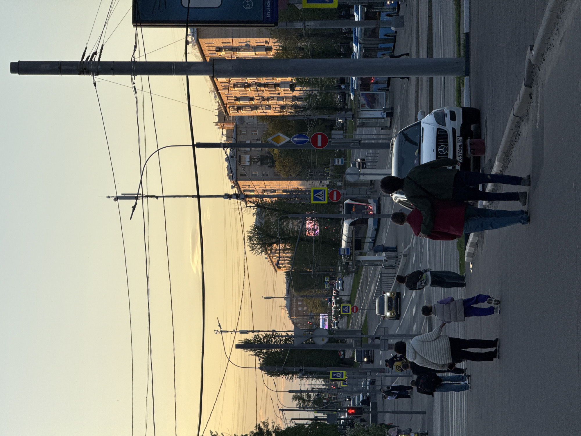 The image shows a street in Moscow, Russia at sunset. The perspective is from a pedestrian's viewpoint on the sidewalk. Several people are walking along the sidewalk. The street is lined with buildings and lampposts. Traffic includes buses and cars. Street signs are visible. A white van with the license plate X574ME37 is parked on the side of the road. Overhead wires run along the street. The sky is a gradient of yellow and orange, suggesting it is dusk or dawn.