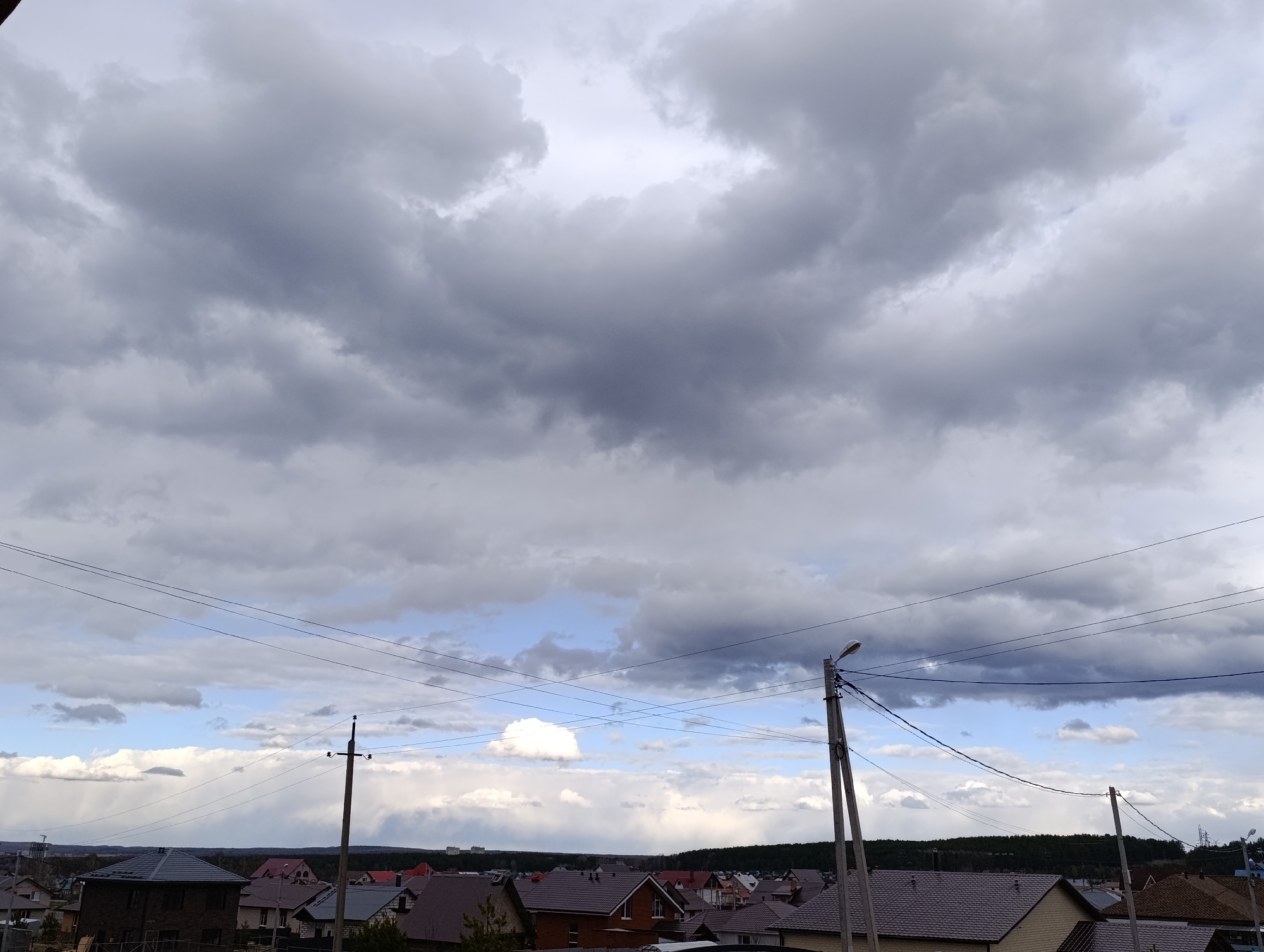 This image captures a broad daytime view of a residential area in Chaikovsky, Russia, dominated by an imposing, heavily overcast sky. Large, dark grey clouds fill the upper portion of the frame, with patches of brighter, lighter blue sky visible lower down and peeking through the cloud cover. The density of the clouds suggests either an impending storm or recently passed inclement weather.
Below the expansive sky, numerous houses with varied, mostly dark-tiled roofs are scattered across the landscape, indicating a suburban or village setting. A distinct line of dense trees or forest marks the distant horizon, behind the clustered homes. Several utility poles, supporting an intricate network of power lines, cut across the mid-ground, providing prominent man-made structures in the scene. One pole in the mid-right features a street lamp. No people are visible, and no specific activities are taking place; the scene is a static landscape. The lighting implies daytime, but the heavy cloud cover creates a diffused, somewhat muted atmosphere. There is no visible text in the image.