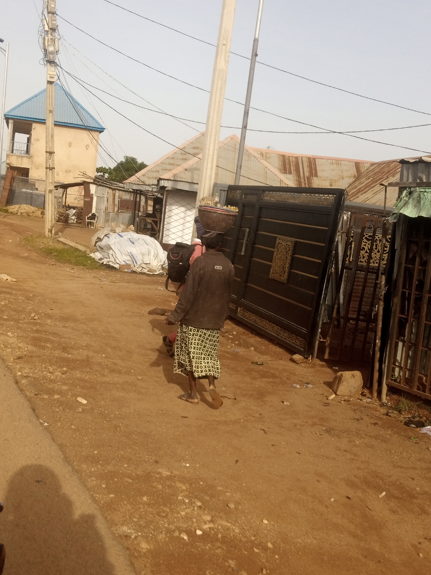 A woman walks along a dirt road, carrying a basket of goods on her head and a backpack on her shoulders. She is wearing a patterned skirt and a dark jacket. In the background, there are several buildings with corrugated metal roofs, and a large black gate. The sky is overcast.
