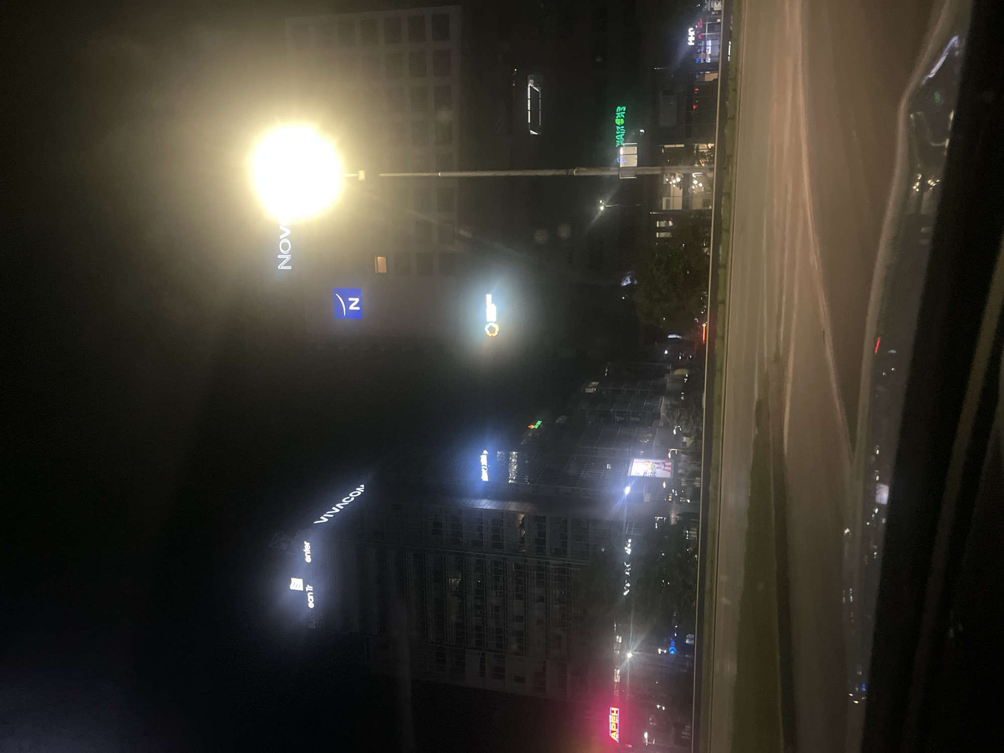 The image depicts a nighttime urban scene, likely taken from inside a vehicle, with a clear view of illuminated buildings and city lights. Prominent building signage indicates the location is Sofia, Bulgaria. The recognizable building signs include "NOVOTEL," and "VIVACOM" are visible. In the background, there is a street with vehicles. The overall environment suggests a bustling urban area at night.