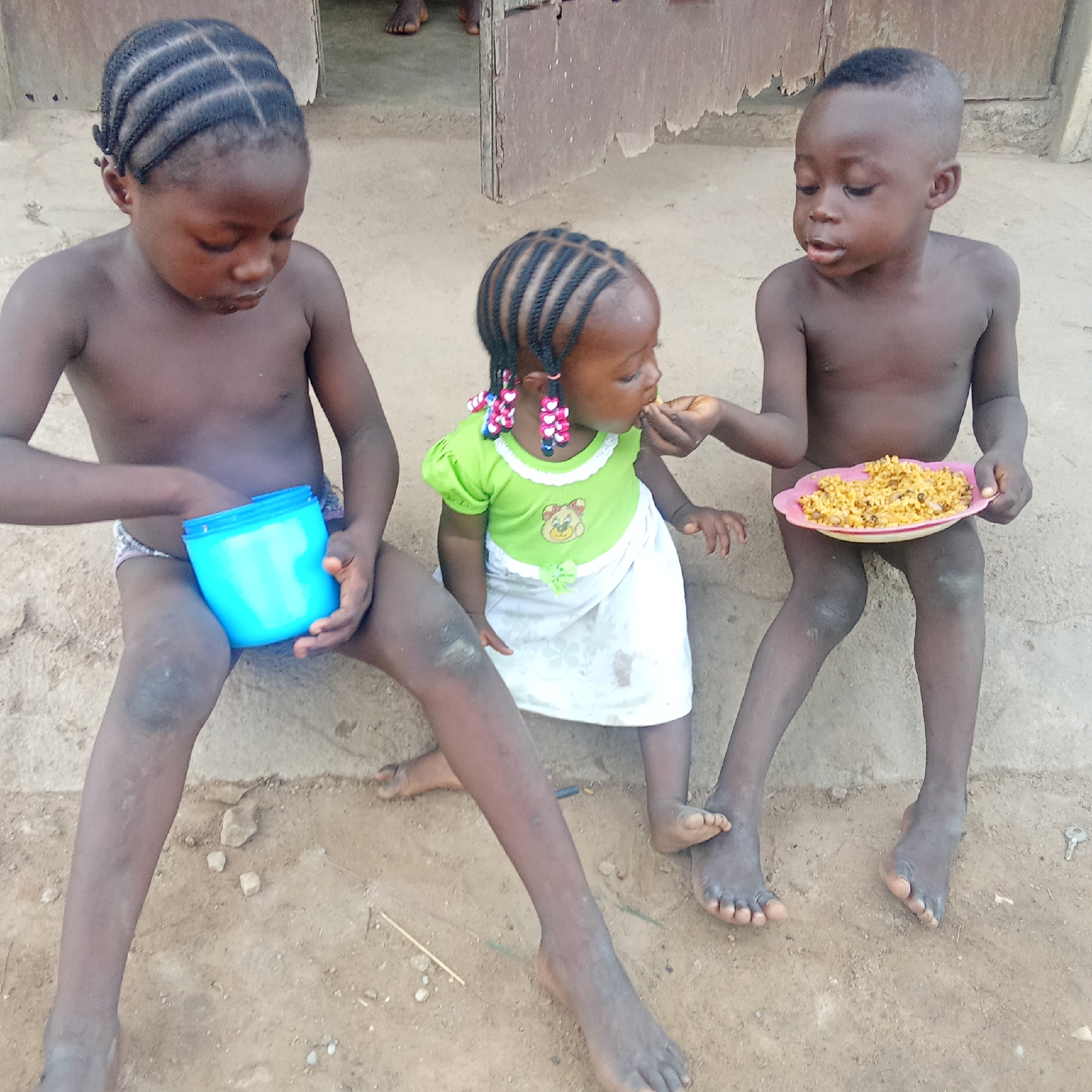 Three young children are sitting on a concrete ledge in Akwanga, Nigeria. Two of the children, a boy and a girl, have cornrow hairstyles. The boy on the right is feeding the girl in the middle from a pink plate of rice or similar food. The girl in the middle is wearing a green shirt with a bear design and a white skirt. The boy on the left is holding a blue container. The ground is dirt. There is a building behind them with a weathered, damaged door.