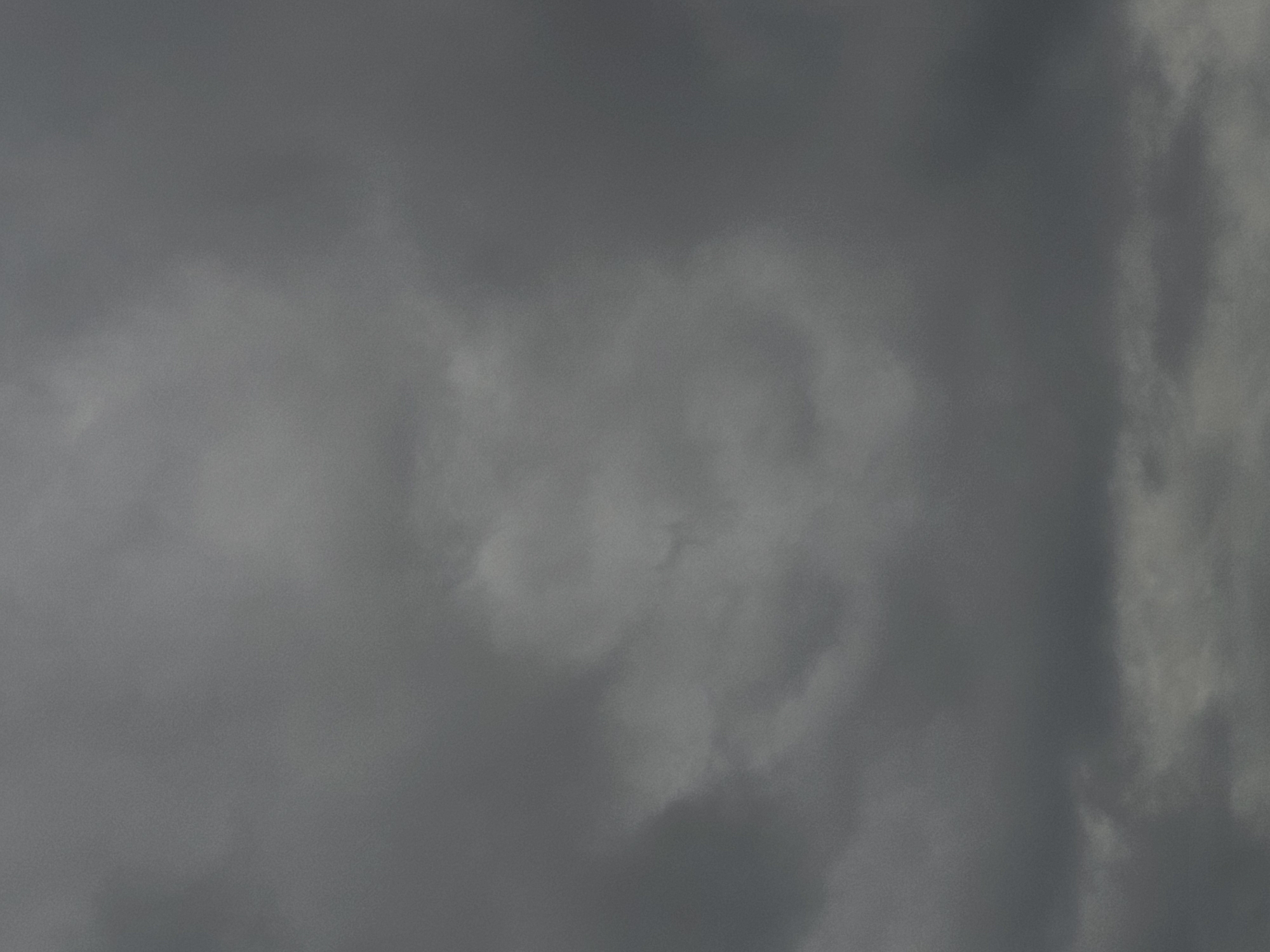 The image depicts a cloudy sky, likely taken during the daytime in Ozolnieki, Latvia. The sky is dominated by dark, gray clouds, suggesting an overcast or potentially stormy weather condition. There are no visible people, subjects, objects, or actions in the frame. The environment is entirely comprised of the sky and its cloud formations, with no other elements of the landscape or cityscape discernible. The lighting is diffuse and even due to the cloud cover, with no strong shadows or highlights. The overall mood of the image is somber and subdued due to the monochromatic color palette and the heavy cloud cover.