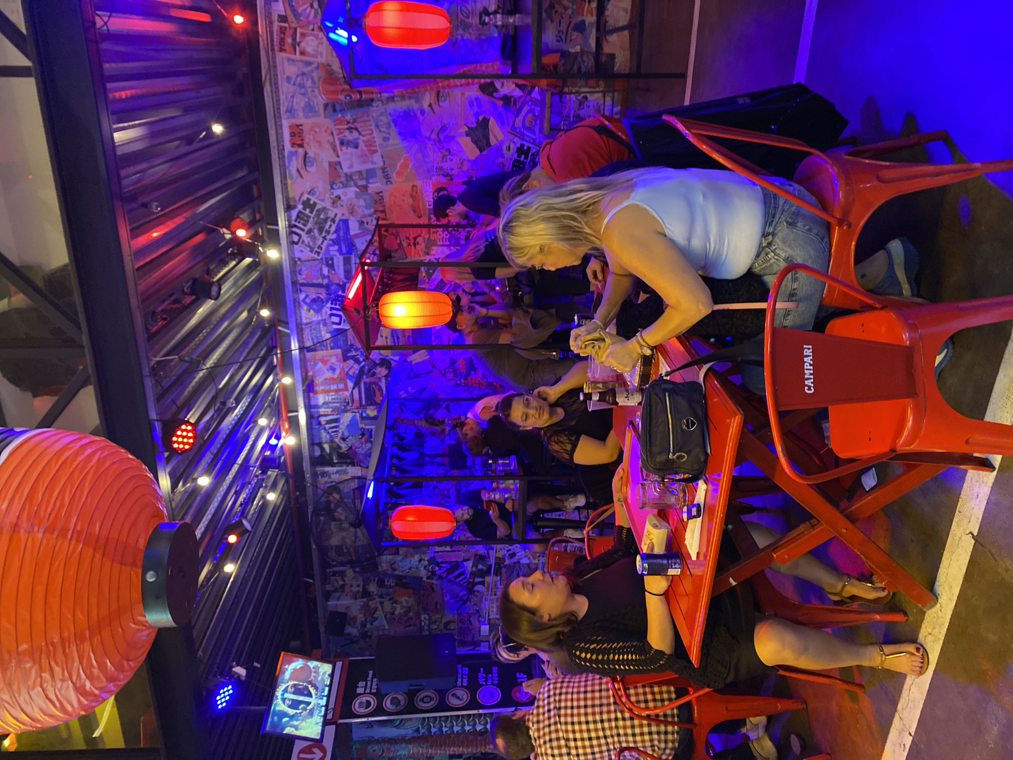 The image depicts an indoor bar or restaurant scene in Buenos Aires, Argentina. A group of people is gathered around red tables and chairs, engaged in conversation and drinking. The walls are decorated with a collage of posters and illuminated with red and blue lighting. A large red lantern hangs from the ceiling. Some of the chairs have the word "CAMPARI" printed on them. One woman is pouring a drink. There is a television screen and a menu of items displayed on the wall.