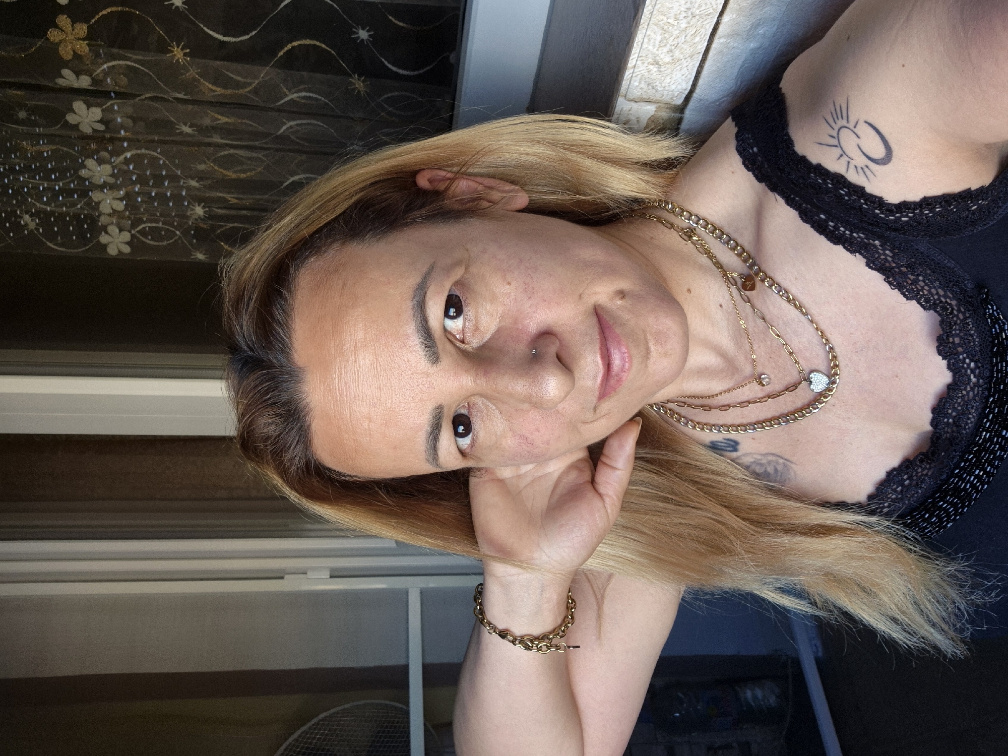A woman with blonde hair and fair skin is pictured in a selfie, with her left arm resting on her head. She is wearing a black top with lace trim and several gold necklaces. A bracelet is visible on her left wrist. A tattoo of a sun and moon symbol is on her right shoulder. The background appears to be a window with sheer curtains and a blurred view of what may be an outdoor setting. The lighting suggests it is daytime.