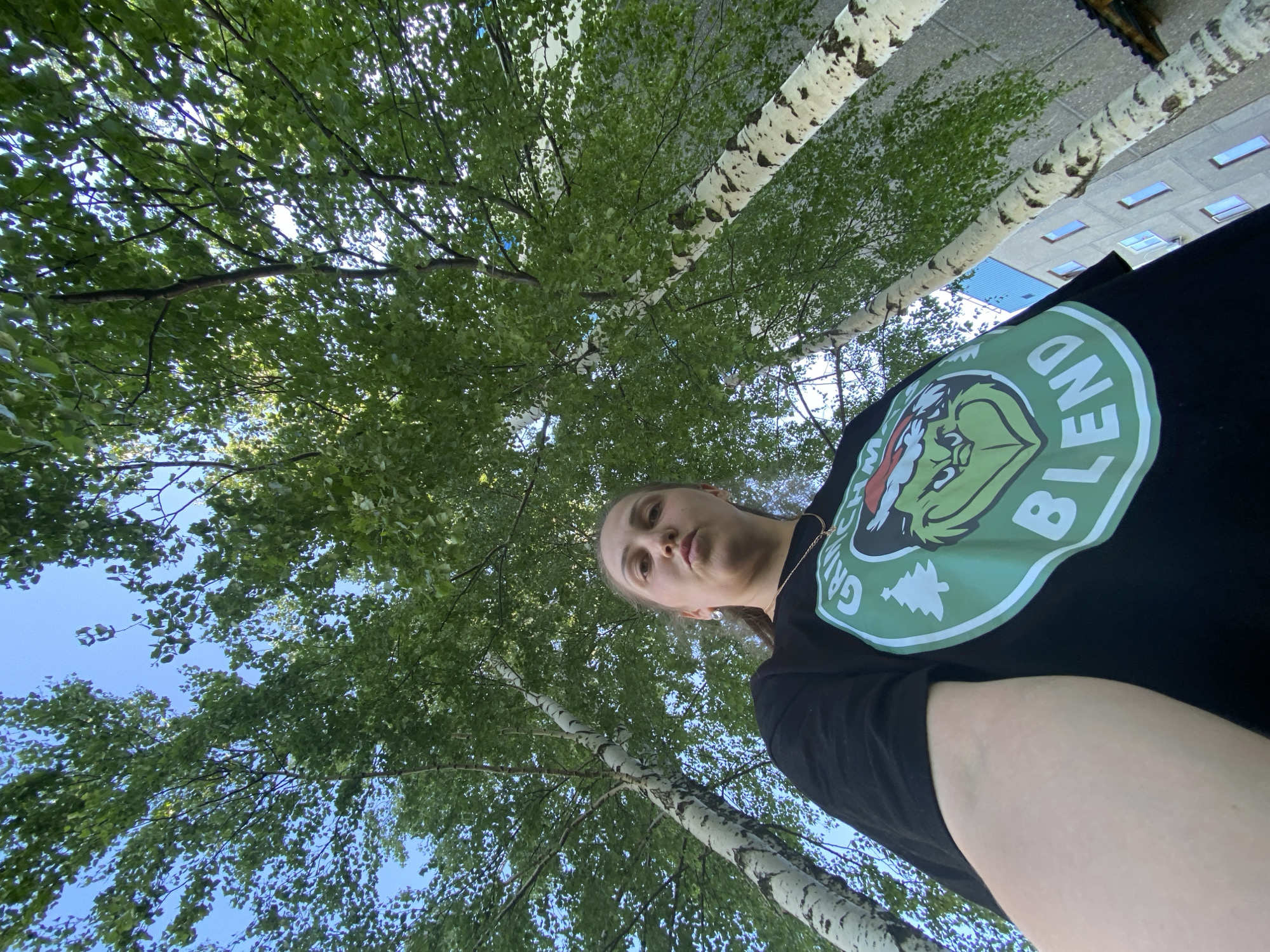A low-angle shot looking upwards reveals a person standing beneath birch trees. The person is wearing a black t-shirt with a green Grinch graphic. Birch tree trunks are visible with mottled white and black bark, surrounded by green leaves. The sky is visible through the leaves. In the top right corner of the image, a building is visible. The location is Марковский, Russia.