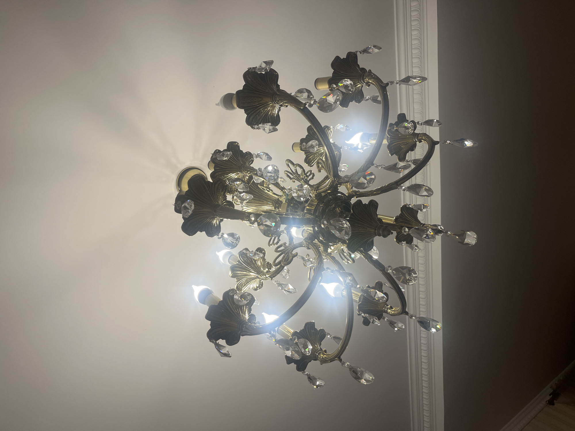 This media file shows a close-up, upward-looking shot of a decorative chandelier. The chandelier is made of what appears to be aged brass, with ornate, leaf-like accents and dangling crystals that catch the light. Several light bulbs are visible, illuminating the fixture and casting light patterns on the white ceiling. The chandelier has multiple arms extending from a central point, creating a multi-tiered effect.
In the background, a white decorative molding or cornice runs along the edge of the ceiling, suggesting an interior setting, likely a room with classical or traditional decor. The lighting suggests it is either nighttime or the room is dimly lit, with the chandelier being the primary light source. The overall mood conveyed is one of elegance and traditional opulence. There is no text visible in the image. The location context is Edem, Russia.