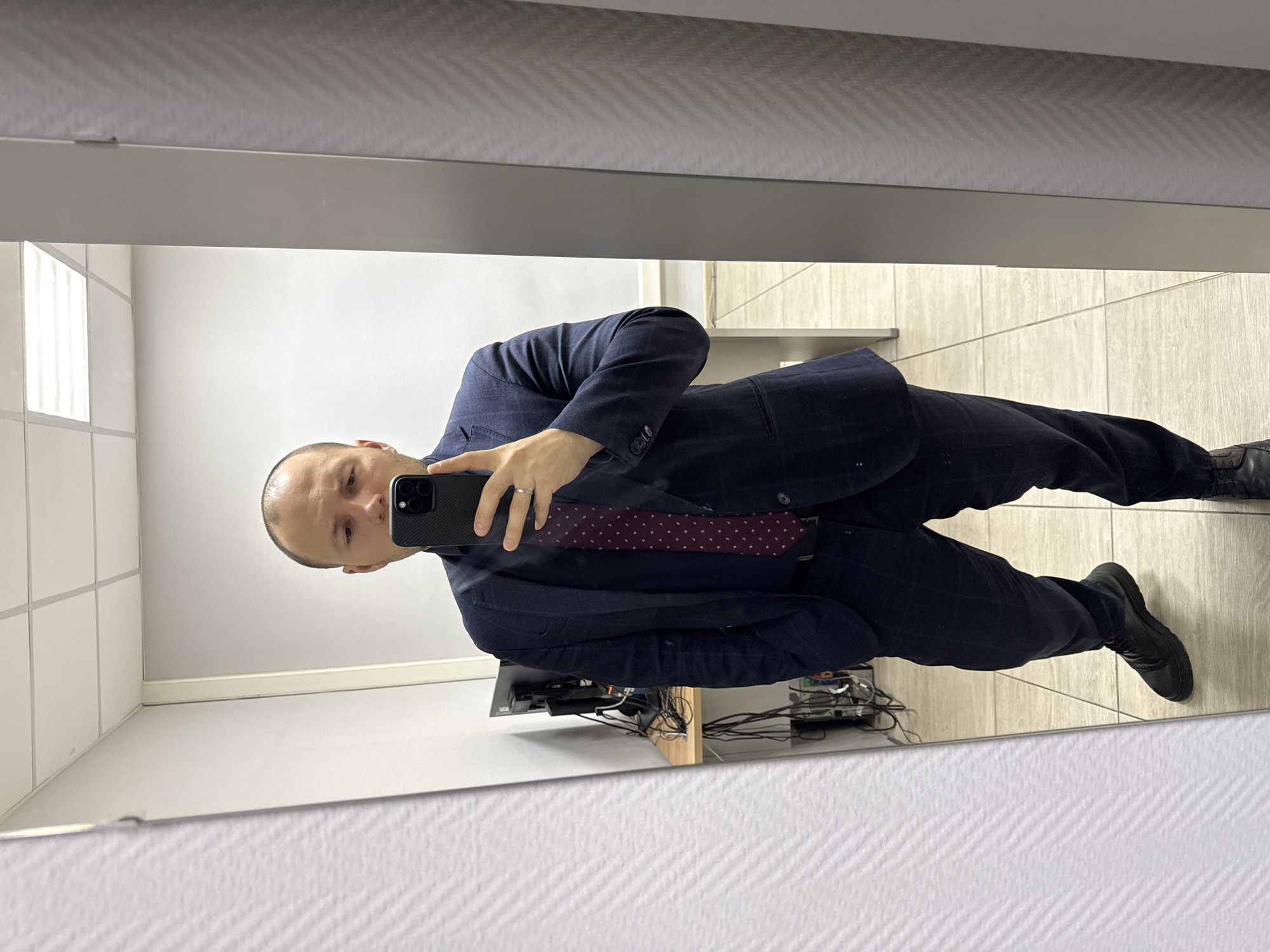 A man is taking a mirror selfie indoors. The man is wearing a navy suit and a burgundy tie with white polka dots. He is holding a black smartphone in his left hand. The floor is tiled, and there's a wall of white tiles to the left of the man in the mirror's reflection. Behind him, there is a desk with electronic equipment and various cables. The location appears to be an office or bathroom, and the city of Krasnoyarsk, Russia is not identifiable from this image alone.