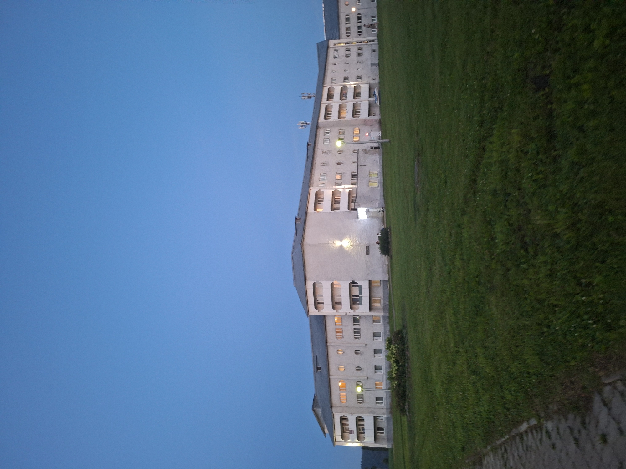 The image depicts a long, multi-story apartment building in the city of Atamanovka, Russia, under a twilight sky. The building is light-colored with a gray roof and features numerous windows, some of which are illuminated, suggesting occupancy. The architecture is functional, with balconies and some circular window elements. In front of the building, there is a well-maintained green lawn with a small landscaped garden bed in the foreground. A paved walkway runs along the left edge of the frame. The scene appears peaceful, with no overt signs of activity beyond a few indistinct figures visible near the building. The overall impression is that of a residential area at dusk.