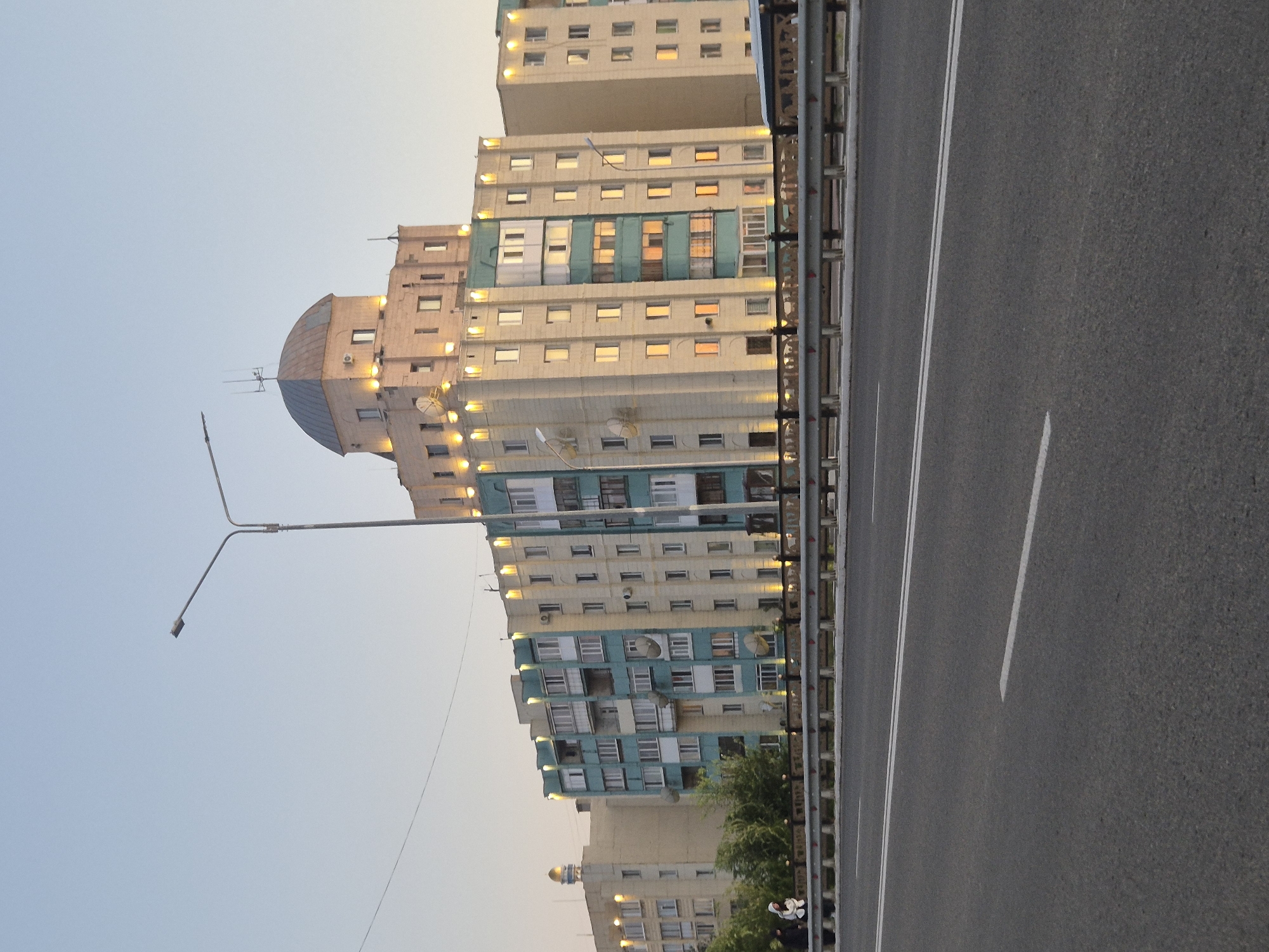 The image depicts a residential apartment building in Kokshetau, Kazakhstan, viewed from an elevated roadway. The building is a multi-story structure with a beige and teal facade, featuring numerous windows and balconies. The top of the building is crowned with a dome-shaped structure, possibly a penthouse or a decorative element, illuminated by external lights. The building is bordered by trees on its left side. A street lamp is visible in the upper left quadrant of the image. The foreground shows a section of an asphalt road with white lane markings. A metal guardrail separates the roadway from the area in front of the building. Two individuals are standing on the sidewalk in front of the building, one wearing a white hooded jacket. The sky is clear and pale blue, suggesting either early morning or late evening light. The overall scene captures a typical urban residential environment.