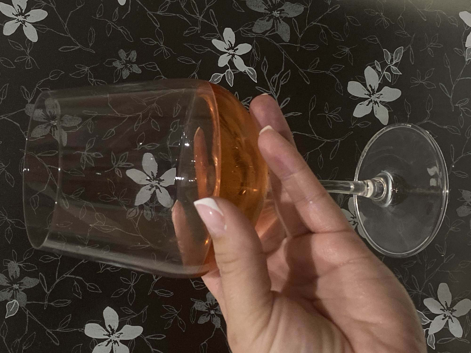 A medium-close-up shot displays a hand holding a wine glass. The wine glass is filled with a transparent orange liquid. The hand has a fair complexion and is adorned with manicured nails featuring a white French tip design. The background consists of a black surface with a pattern of stylized white flowers and branching stems. The floral pattern appears to be printed or woven into the surface. The precise location within Sochi, Russia, cannot be ascertained from the provided image.