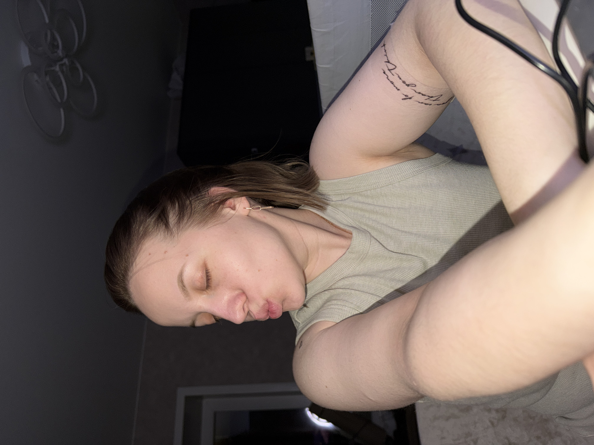 A young woman is reclining, possibly lying on a bed or other soft surface. She is wearing a light green or gray tank top. Her left arm is raised and slightly bent, revealing a tattoo on her upper arm, and a black cable is visible partially obscuring her arm. Her eyes are closed, and her lips are pursed. The background consists of a dimly lit room with a dark-colored wall. A decorative white object is mounted on the wall, and a TV or monitor is partially visible. In the bottom part of the image, a dimly lit door or window is also visible. The location is not detectable from this image.