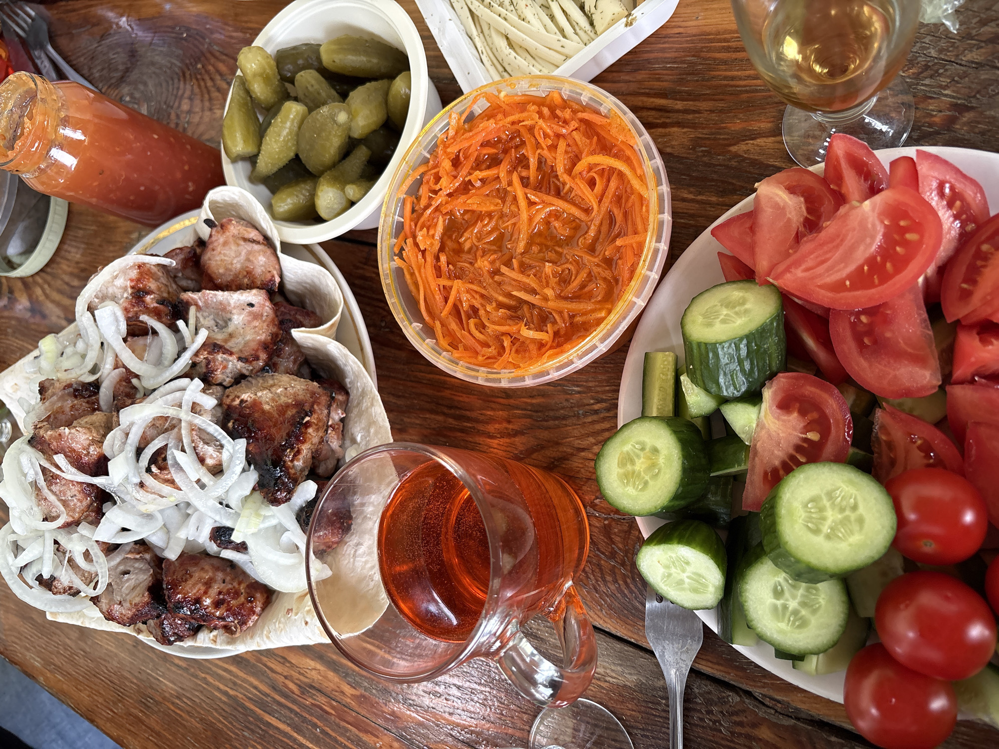 A wooden table is laden with food and drinks. From left to right, there is a plate of shish kebab topped with sliced onions, a jar of red sauce, a container of pickled gherkins, a container of shredded carrot salad, a container of string cheese, a glass of white wine, a plate of sliced tomatoes and cucumbers, and a glass mug filled with an orange-colored liquid. The location is Dubovaya, Russia. No people are visible in the image.