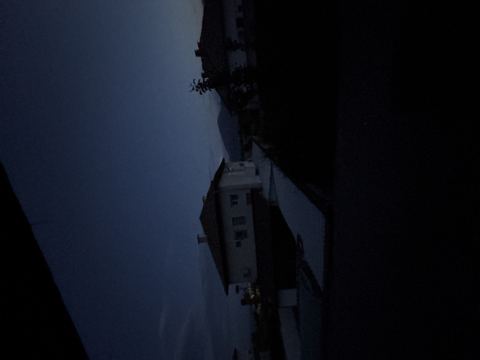 The image depicts a serene twilight or nighttime scene, likely in Garmen, Bulgaria. The original image is rotated 90 degrees counter-clockwise, but when viewed correctly, it reveals a quiet landscape under a dark sky.
A prominent subject is a light-colored, multi-story house situated towards the left of the frame. It features a reddish-brown roof, multiple visible windows, and a chimney. Behind the house and extending towards the right, the dark silhouettes of trees or bushes with discernible branches stand out. In the distant background, faint outlines of dark mountains or hills are visible against the deep blue, almost black, evening sky. The sky dominates the upper portion of the image. The foreground and the right side of the scene consist of indistinct, shadowed ground or terrain.
The time of day is clearly evening or night, indicated by the very low light conditions and the dark, deep blue hue of the sky. There is no apparent artificial lighting from the house or within the landscape. No specific activity or event is taking place, and no people are visible. There is no discernible text within the image. The overall impression is one of tranquility and quietude in a rural or suburban setting.