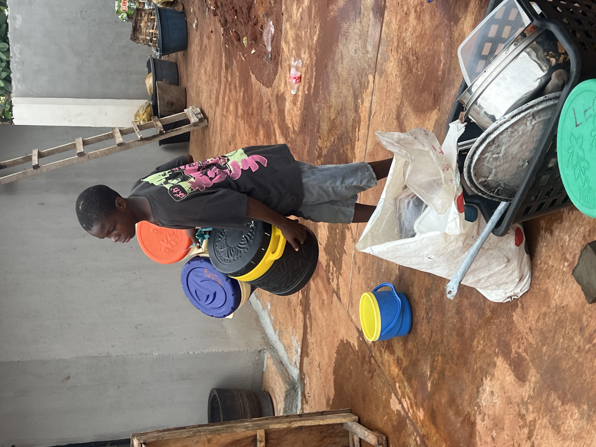 The image captures a young boy engaged in an activity within an outdoor or semi-outdoor setting in Onitsha, Nigeria. The boy, of African descent, wears a dark t-shirt with a vibrant graphic design featuring pink and green elements, including visible Japanese characters that appear to say "がんば" (ganba, likely part of "Ganbare!" meaning "Do your best!"). He is also wearing light-colored shorts.
The scene takes place on a reddish-brown ground, which appears damp or wet in various spots, reflecting light. A light gray concrete wall is visible to the left, with some green foliage beyond it. In the background, a wooden ladder leans against the wall, and several black plastic tubs are present, one seemingly filled with dried vegetation or wood.
In the foreground, the boy is surrounded by several colorful plastic containers and lids. He is looking down at a stack of these items, including an orange lid, a blue lid, and a black container with a yellow handle, on which "SUN..." is faintly visible. To his right, a small blue and yellow bucket sits on the ground. Next to the bucket, a large white bag, possibly filled with items, leans against a black plastic basket. This basket contains various household objects, such as shiny metal bowls or pots, a clear plastic container, and a green plastic lid with a decorative pattern, on which "LG" is partially visible. A red plastic bottle lies on the ground nearby.
The boy appears focused on the containers, possibly sorting them, playing, or preparing to carry them. The overall impression is of a daily domestic activity in a functional, possibly somewhat cluttered, outdoor space during daylight hours. No specific time of day or weather is explicitly indicated beyond the general brightness and damp ground.