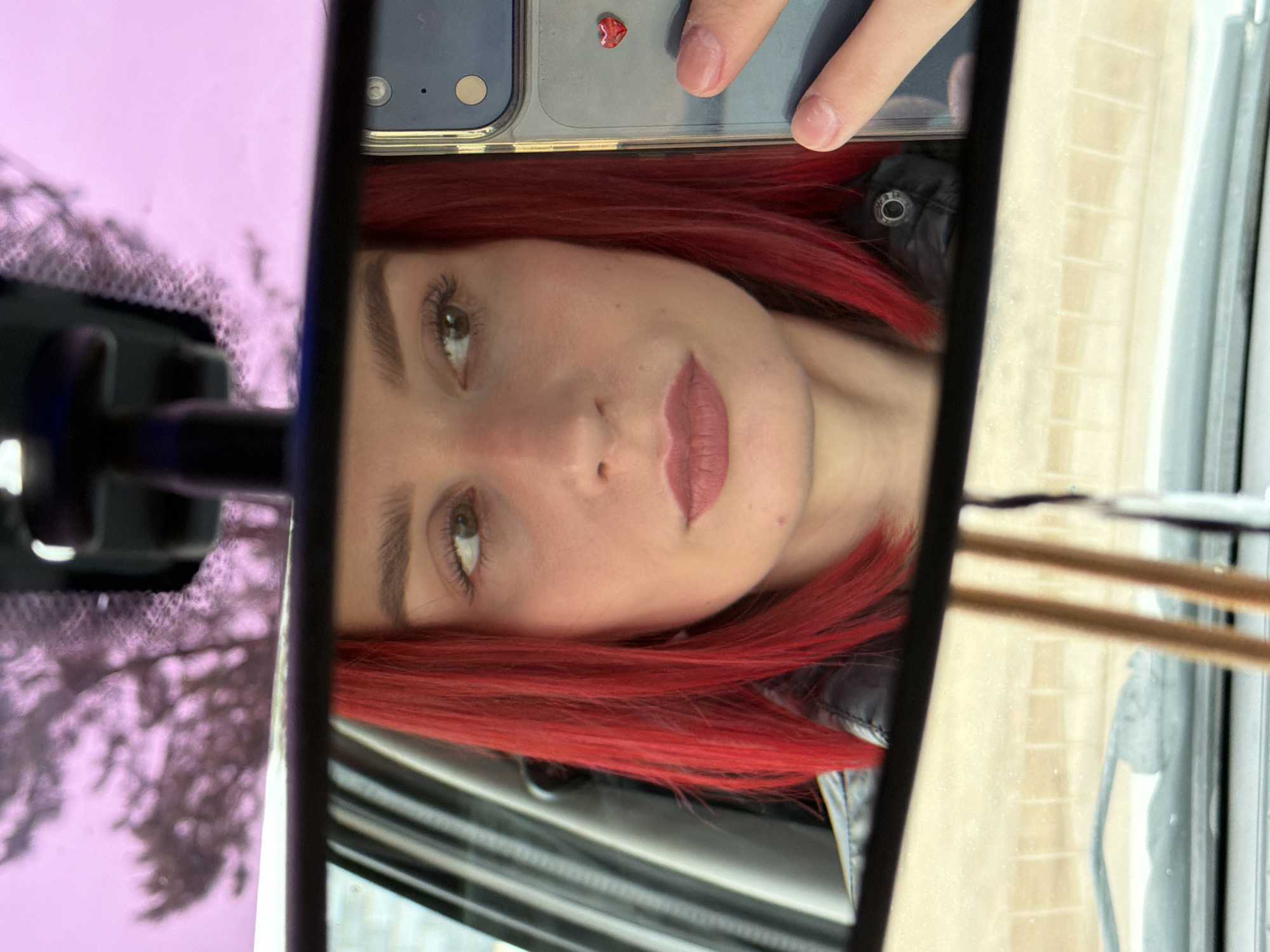 A selfie captures a woman with red hair, reflected in what appears to be a car's rearview mirror. The woman's face is centrally positioned. The rearview mirror frame is visible on the left and right, showing glimpses of the vehicle's interior and surroundings. A smartphone is visible at the top of the frame, held above the mirror. The overall composition suggests the photograph was taken inside a car, possibly in Chaikovsky, Russia, although the exact location is not discernible within the frame itself.