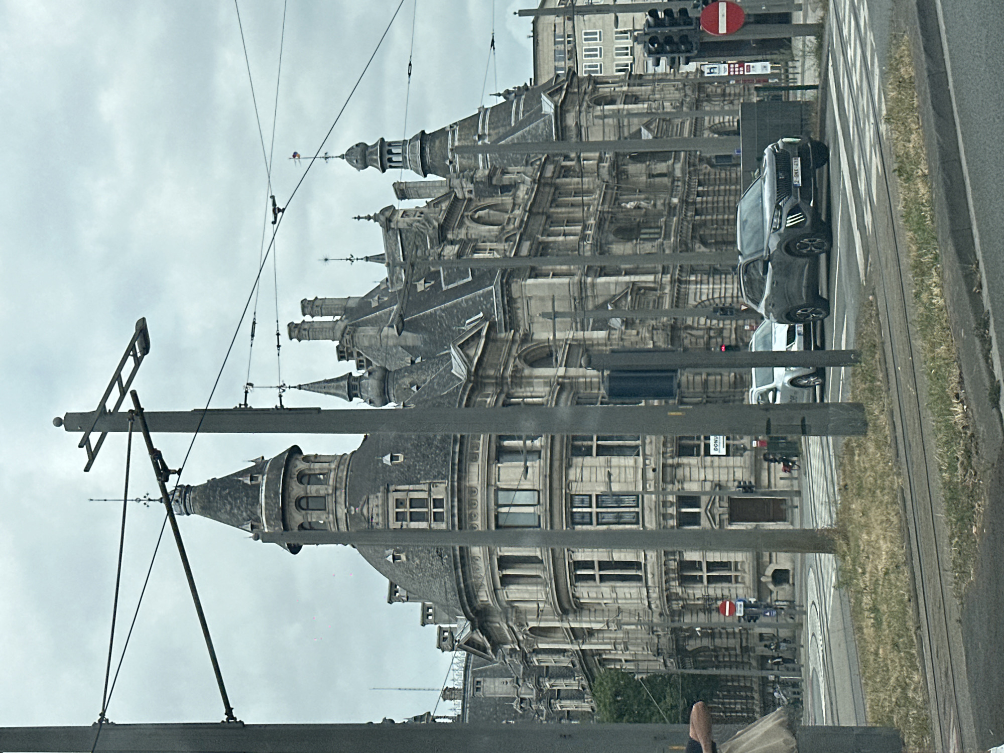 The image displays a street scene in Antwerp, Belgium, characterized by historic architecture and urban elements. The perspective is from a moving vehicle, likely a tram or car, as indicated by the blurry foreground and the presence of overhead wires for a tram system. The buildings are ornate, featuring classical architectural styles with detailed facades, arched windows, and prominent rooflines with spires and turrets.
A dark-colored SUV is visible on the right side of the road, stopped at a traffic light. The license plate is partially obscured but appears to be European. Other cars and elements of urban infrastructure, such as traffic lights and crosswalks, are also present. The sky is overcast, suggesting a cloudy or possibly rainy day. The vegetation along the roadside is green and somewhat unkempt, hinting at a temperate climate.
The composition is dominated by the grand buildings and the overhead tram wires, which create a layered effect. The image is oriented vertically, and the reflection of the scene in a glass surface is also visible, adding another dimension to the visual information. There is no explicit indication of people engaged in specific activities, though some figures are visible in the distance, suggesting general pedestrian traffic. The overall mood is one of everyday urban life in a city with a rich architectural heritage. The time of day appears to be daytime, given the ambient light.