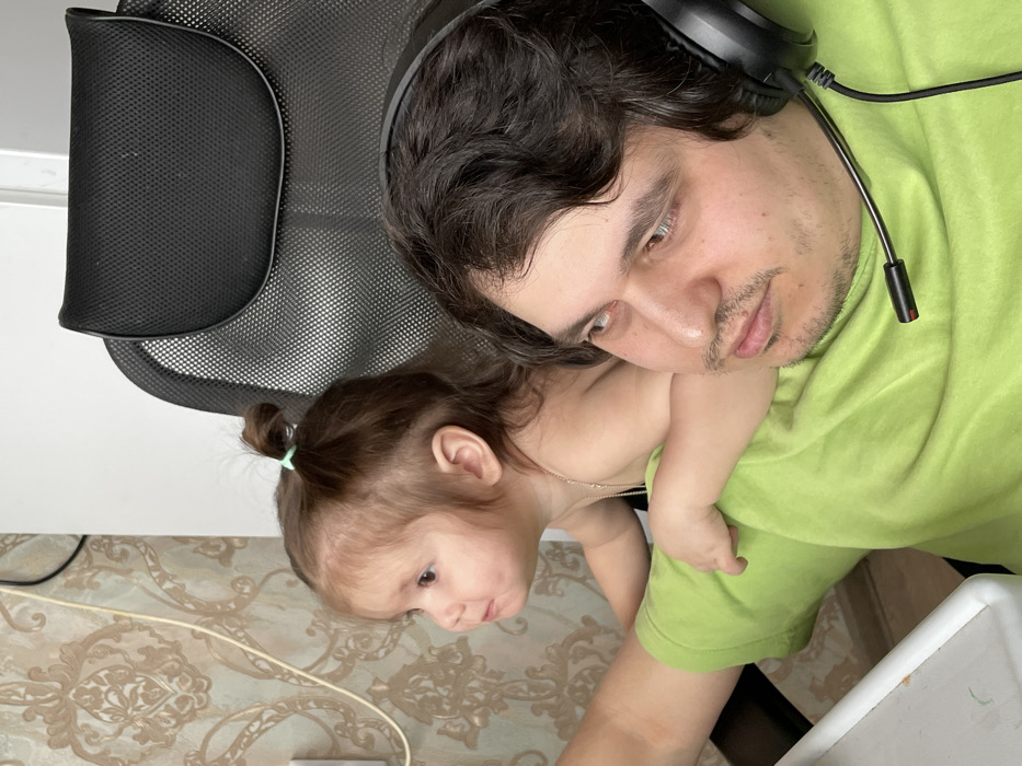 An indoor scene depicts two individuals, an adult male and a young child, positioned closely together. The adult male, with dark, wavy hair and a light mustache and goatee, wears a lime green short-sleeved shirt and black over-ear headphones with an attached microphone boom. His gaze is directed to the observer's right, appearing focused. A young child, with light brown hair tied in a small ponytail with a light blue elastic, is positioned behind the male, leaning over his right shoulder and upper back. The child is bare-chested and wears a delicate, light-colored beaded necklace. The child's right arm rests on the male's upper right arm. The child's gaze is directed to the observer's left. In the background, patterned wallpaper featuring a light brown and beige damask or floral design is visible. A white electrical outlet and associated cables are present on the wall. A portion of a black mesh office chair's headrest is visible behind the child. A white surface, possibly a desk or tray, is partially visible in the lower left corner. The image lacks specific visual cues to determine its location within the city of Izhevsk, Russia.