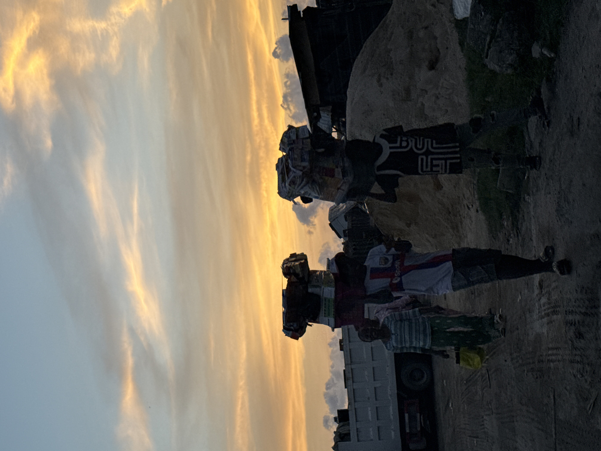 In Ado, Nigeria, the image captures three individuals near a truck under a sky tinted with orange hues, likely during sunrise or sunset. The people stand adjacent to what appears to be a construction site or waste disposal area with sand and scattered debris. The individuals are dressed in casual clothing, with one wearing a shirt branded with "Spotify" and another with "BULLET" on it. The truck is parked nearby, suggesting their involvement in loading or unloading materials.