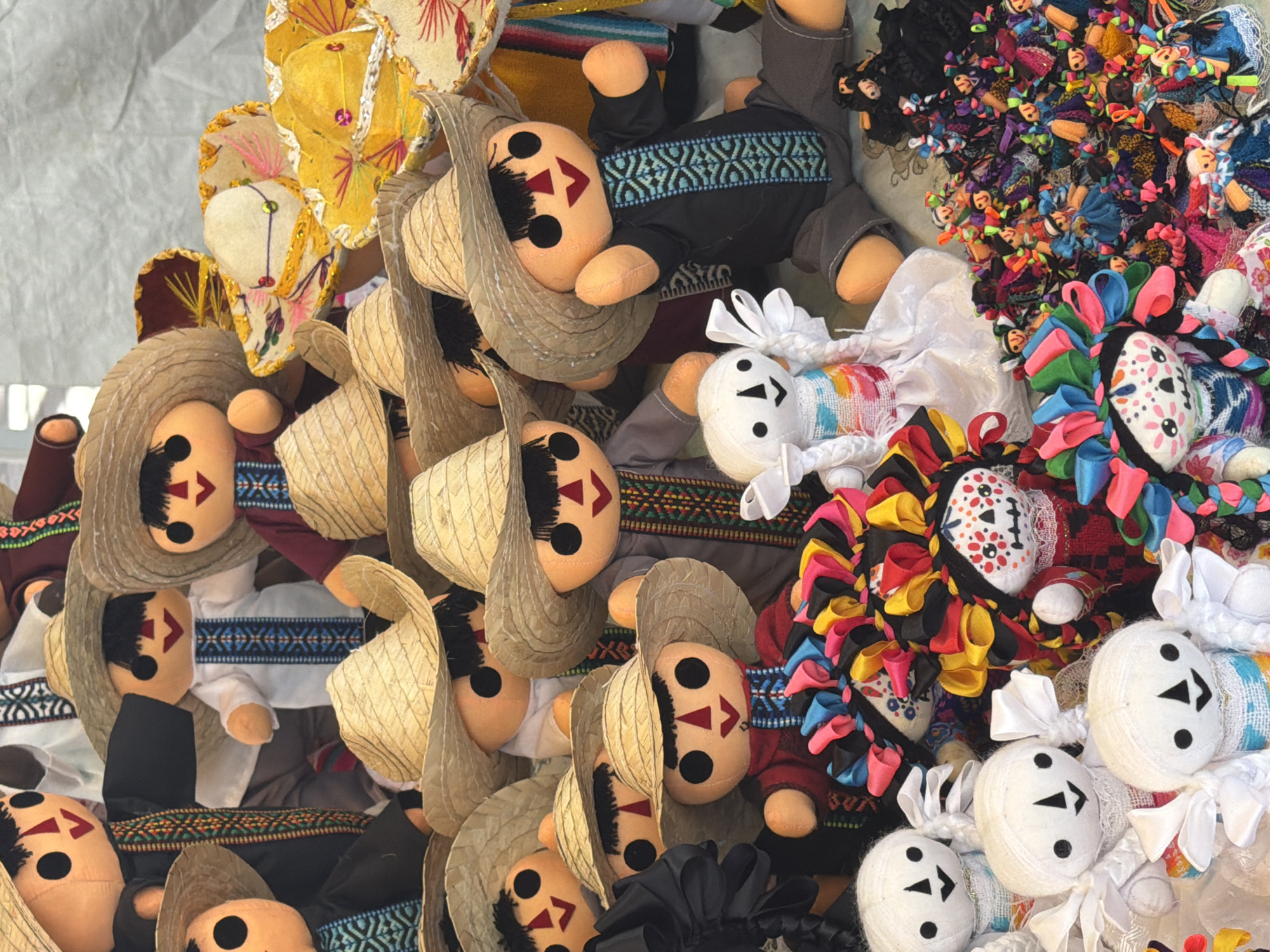 The image presents a high-angle, close-up view of a collection of handcrafted dolls, possibly for sale at a market. The dolls exhibit various designs, including those with straw sombreros, those dressed in black embroidered shirts, and those adorned with colorful ribbons or styled as Catrina dolls with skull faces. The scene appears to be a display of cultural or tourist items, typical of a Mexican craft market. The dolls are densely packed, indicating a bulk presentation strategy. The background is nondescript. There is no specific mention of the location.