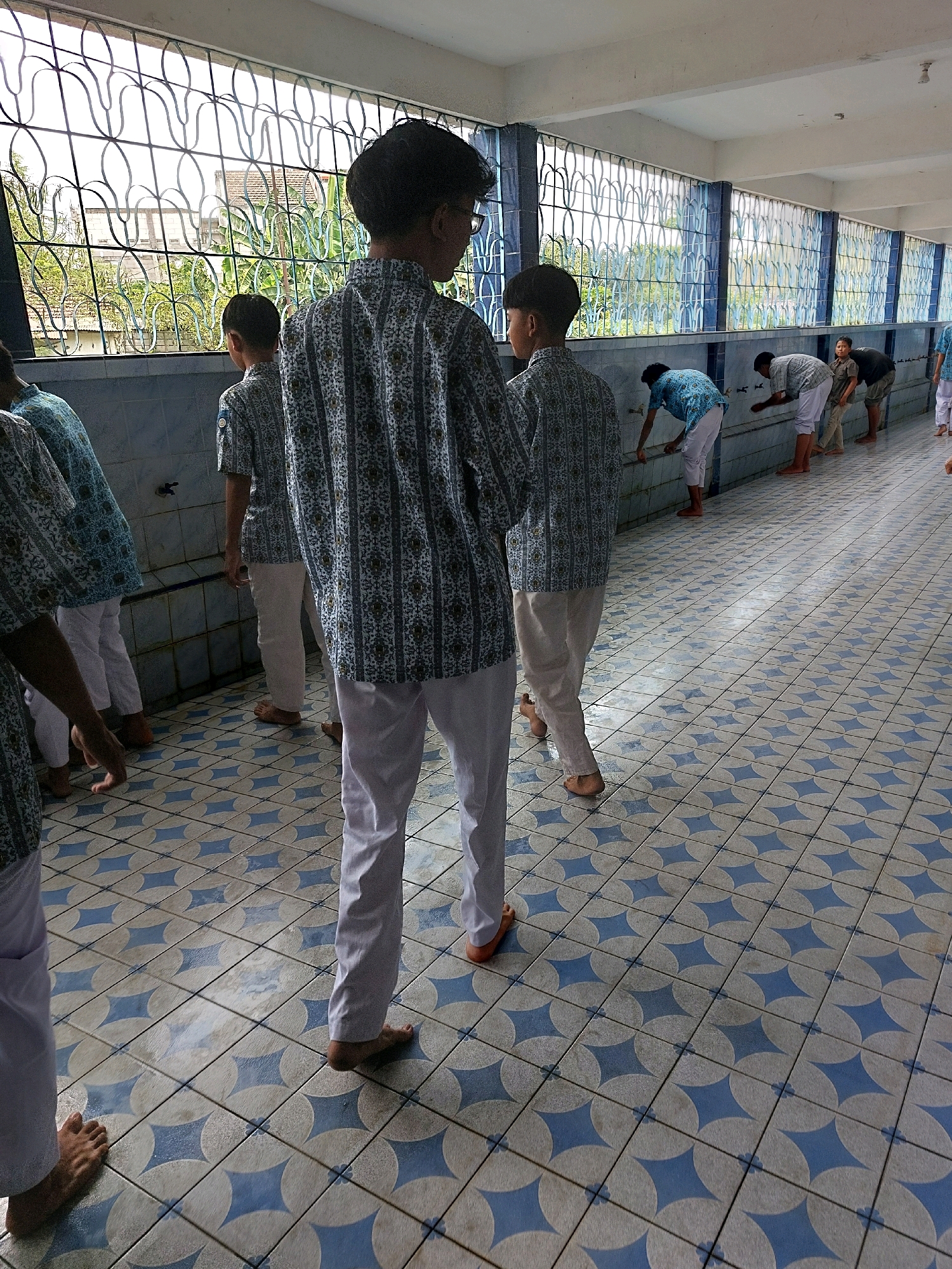 In the city of Sumberrejo, Indonesia, a group of boys in school uniforms stand in a tiled hallway with a patterned floor, which is made up of light beige and blue diamond shapes. They are standing at a sink area with blue tiled walls and a metal faucet. Outside a large window, greenery and buildings are visible.