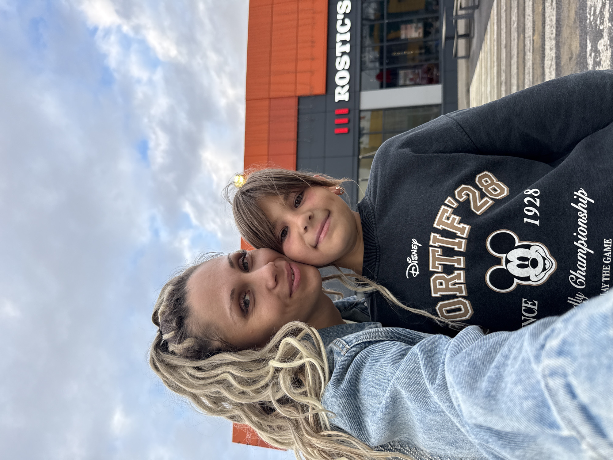 A mother and daughter are taking a selfie outdoors in front of a building with orange and dark grey panels. The building has a sign that reads "ROSTIC'S". The mother is on the left side of the frame, wearing a light blue denim jacket. She has long, blonde, curly hair pulled back in a high ponytail. The daughter is on the right side of the frame, wearing a dark grey sweatshirt with a Disney graphic featuring Mickey Mouse and the text "SPORTIF 28 1928". She has brown hair and is smiling. They are standing in front of a crosswalk. The sky above them is cloudy. The location is identified as Ignatovo, Russia.