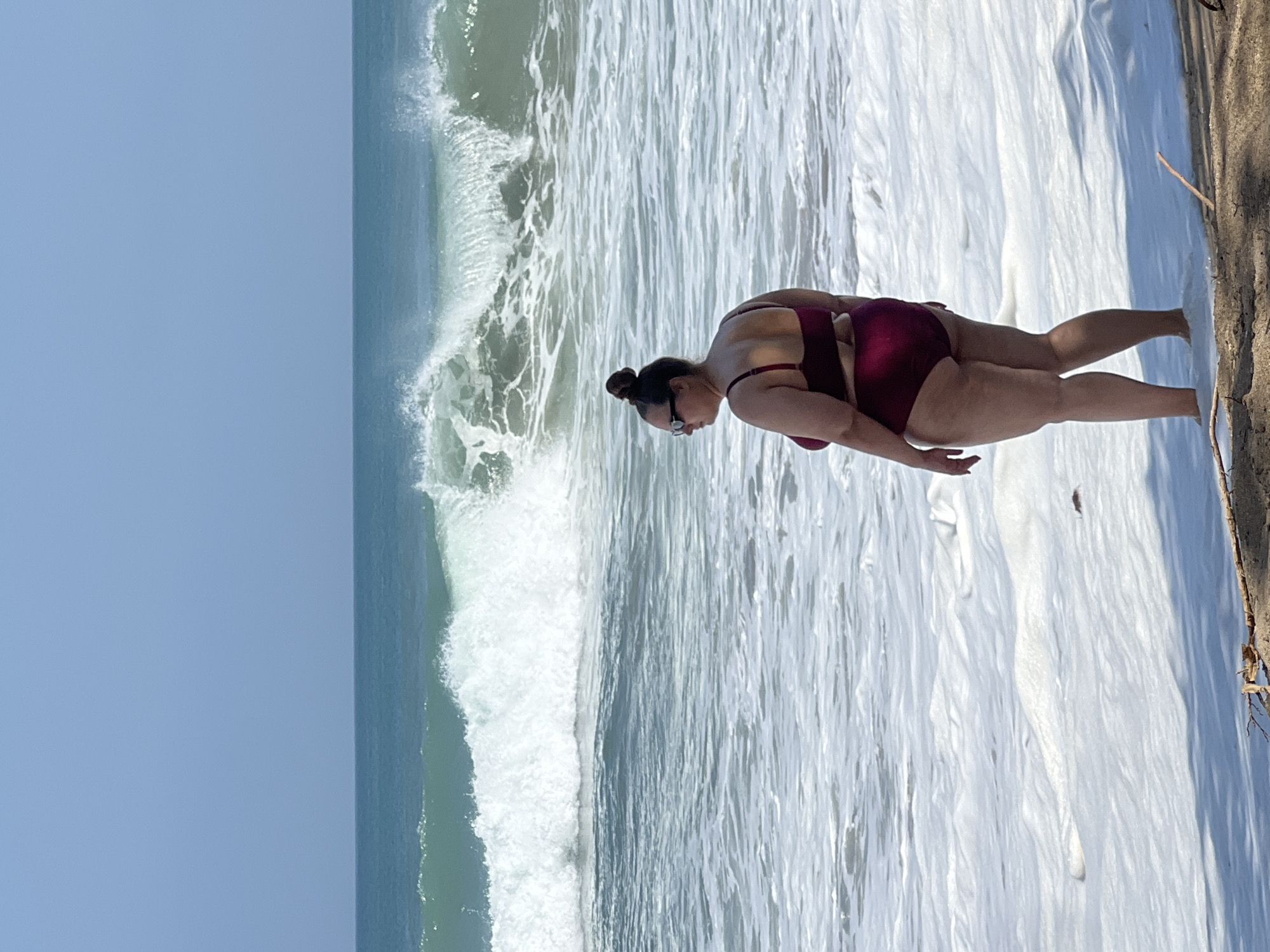 A full-body, medium-shot photograph captures a person standing at the edge of the water on a beach in Kuta, Indonesia. The individual, facing away from the camera and to the left, is wearing a maroon bikini. Their hair is styled in a bun on top of their head, and they are wearing sunglasses. The ocean in the background features a large, breaking wave with white foam. The sky is a clear, pale blue. The foreground is a sandy beach with some shadows cast from vegetation. The image appears to have been taken in daylight, likely during the day.
