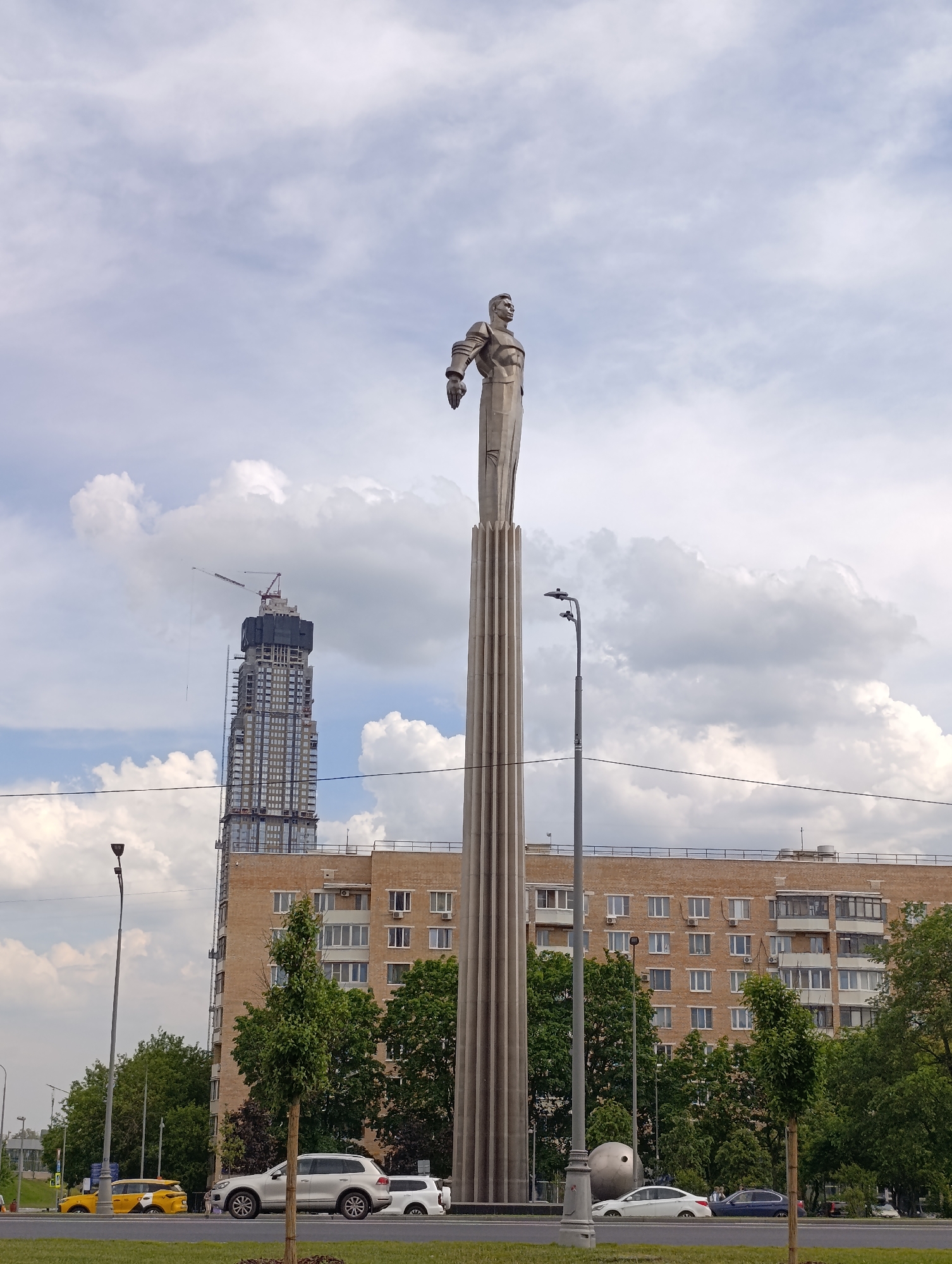 This eye-level, vertical shot captures an urban scene in Moscow, Russia, under a bright, partly cloudy sky during daytime. The central subject is a towering monument featuring a heroic, space-themed figure standing atop a tall, fluted, rocket-like pedestal. The figure, depicted in a stylized uniform with one arm slightly raised, gazes skyward.
Adjacent to the monument's base, on the left side of the frame, a large, metallic spherical sculpture, resembling a satellite (likely Sputnik), is visible. Several cars, including white SUVs and yellow taxis, are parked or driving on the road in the foreground.
In the mid-ground, a multi-story, light-brown brick building with numerous windows and balconies provides a backdrop, partially obscured by green trees. To the left and behind the brick building, a very tall, modern skyscraper is prominently under construction, topped with a crane. A streetlight stands to the right of the monument.
The scene combines monumental public art with contemporary urban development and daily street life, characteristic of a bustling city. The weather is clear with scattered clouds, and no discernible text is visible.