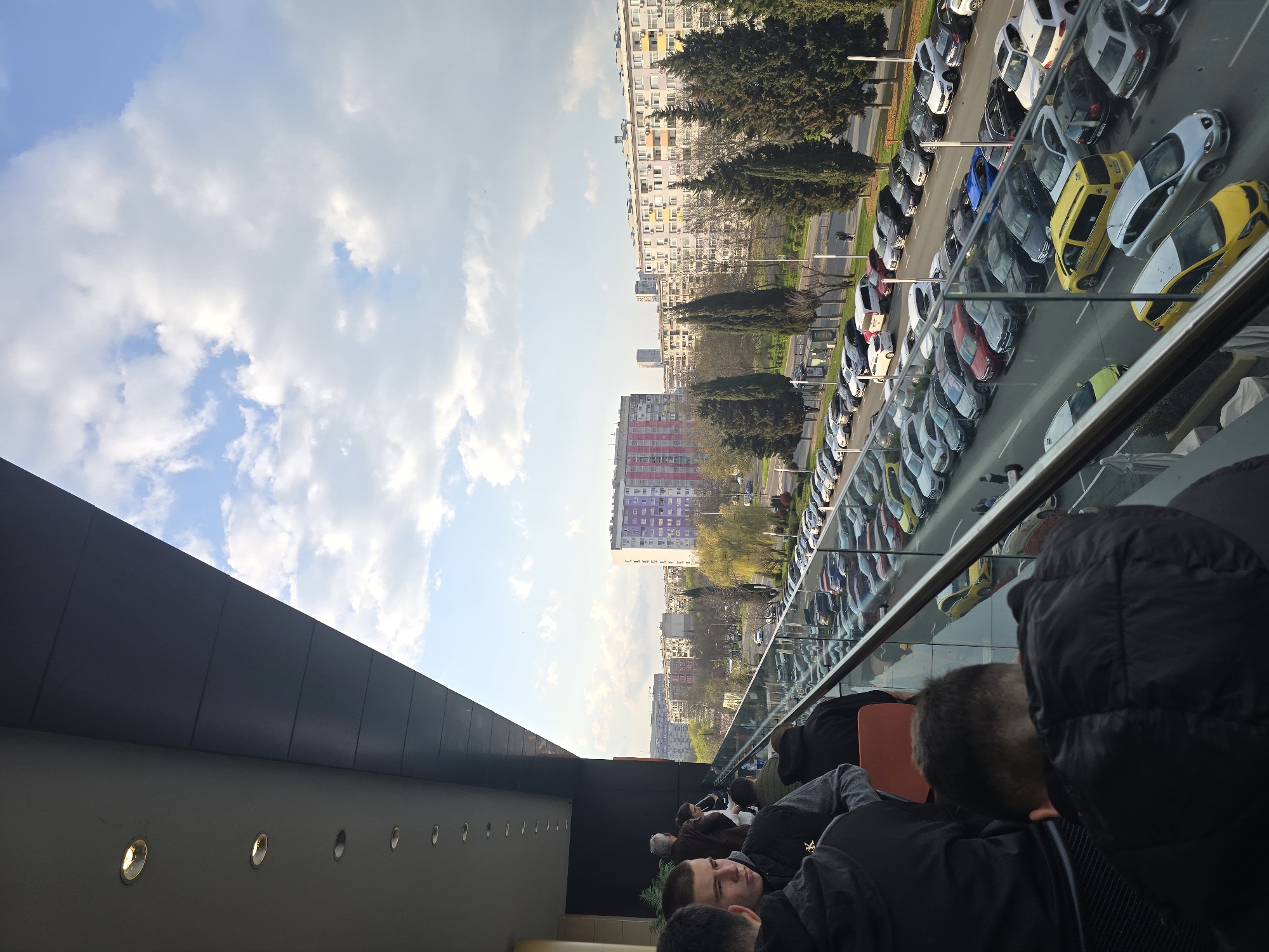 From an elevated perspective, possibly a building or structure with a balcony, a row of parked cars is visible alongside a street. Buildings are seen in the background, characteristic of an urban setting. People are present on the balcony, some seated, seemingly observing the scene below. The sky is partly cloudy, and the lighting suggests daytime. The general location is identified as Burgas, Bulgaria, based on recognizable architectural and environmental characteristics of the city.