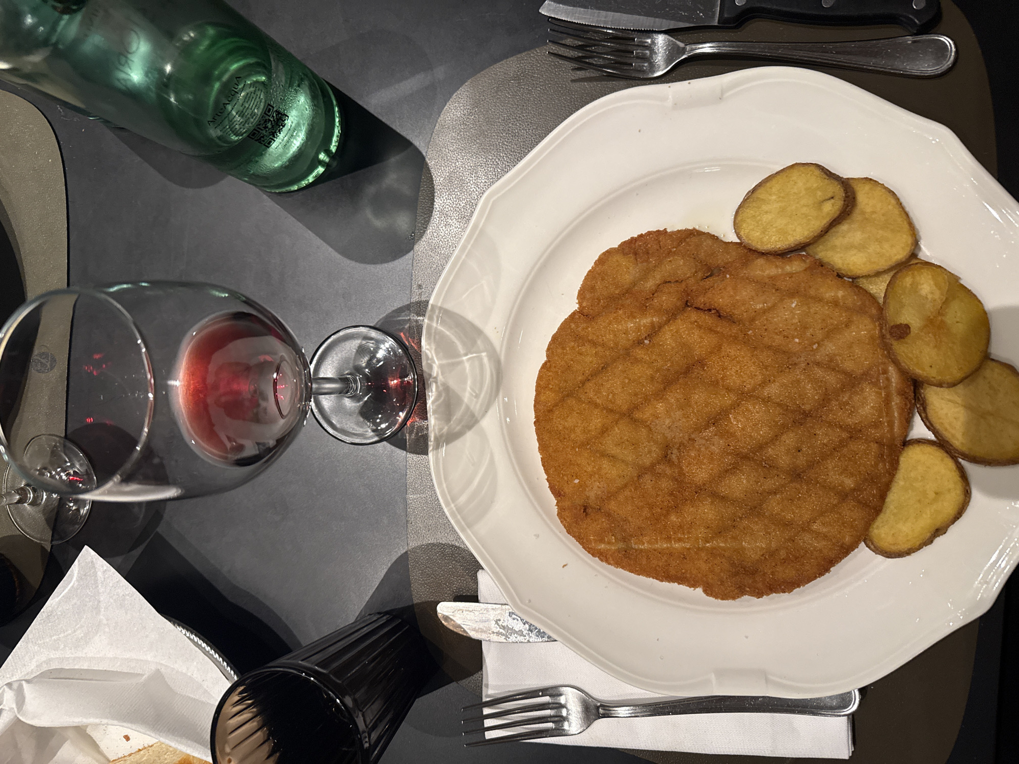 The image depicts a restaurant table setting, likely in Milan, Italy, based on the typical presentation of the meal. A white, fluted-edged plate dominates the scene, holding a large, breaded cutlet (likely a Milanese cutlet) with fried potato slices arranged along one side. To the left of the plate is a wine glass filled with red wine, and next to it is a partially visible green glass bottle. Further to the left, partially visible, is a bread basket and a drinking glass. The table is dark, with light reflecting off of it, with cutlery arranged on the table around the plate. A napkin is placed underneath the fork and knife. The photo is taken from an overhead perspective.