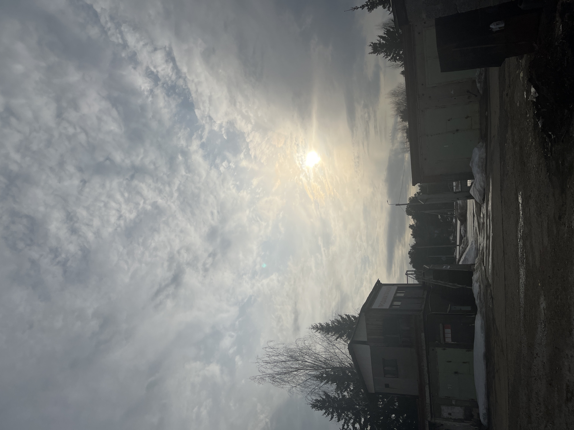 The image depicts a daytime scene in what appears to be an industrial or semi-urban area, likely in Chaikovsky, Russia. The sky is overcast with high, thin clouds, and the sun is visible but obscured by the cloud cover. The foreground consists of a dirt or gravel road, slightly wet or muddy. To the right, there's a row of dark green garage-like structures. Further in the background, there's a building, possibly two-storied, also appearing industrial. Tall trees, likely evergreen, are scattered throughout. The lighting is flat and muted. There is some snow on the ground in patches. No people are visible in the scene.