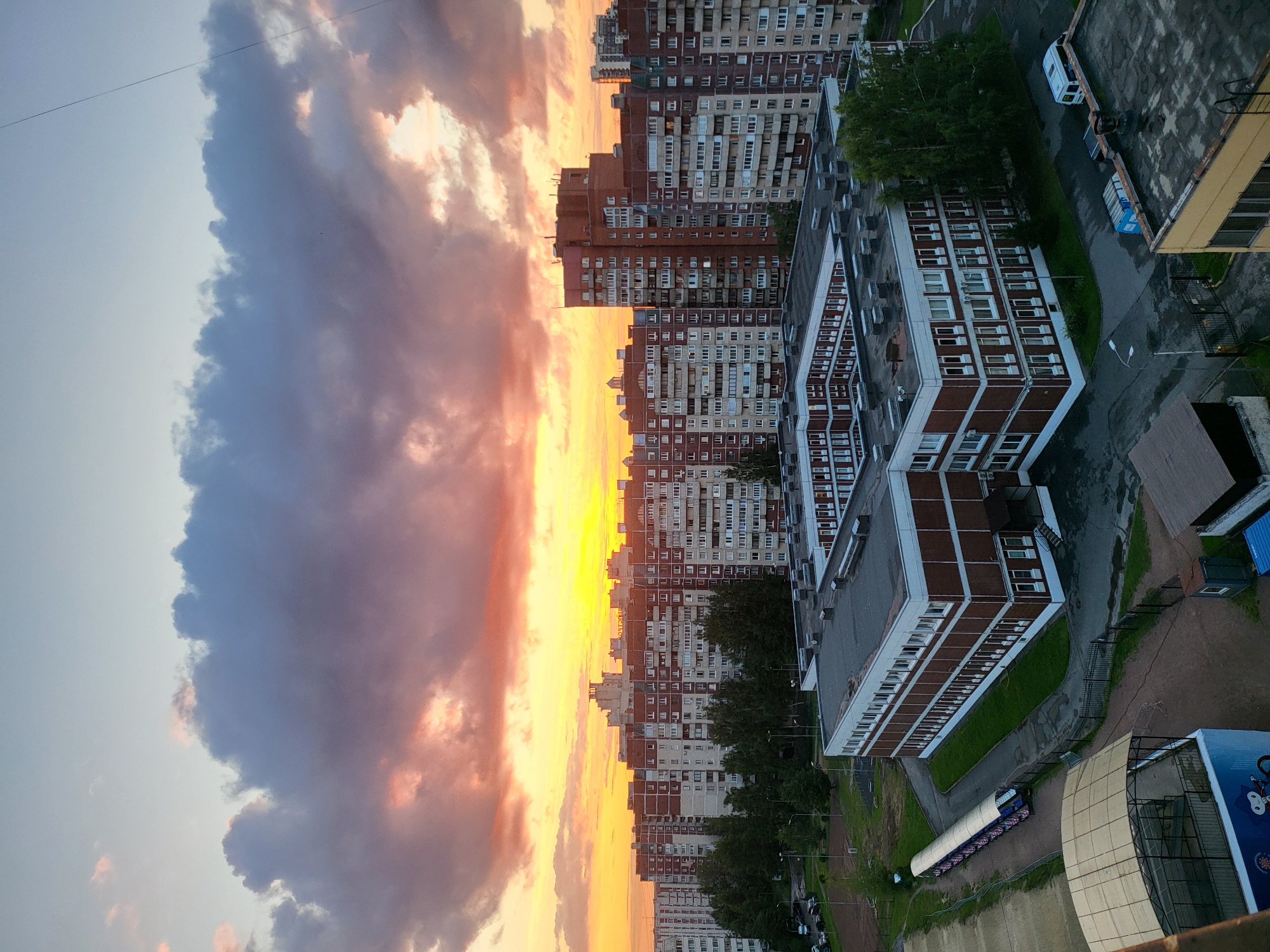 This is an aerial view of a residential area in Saint Petersburg, Russia, during sunset. The sky is filled with dramatic clouds, colored orange, pink, and purple by the setting sun. In the foreground, a modern, U-shaped, multi-story building with a red brick facade and white window frames is visible. This building appears to be a school or educational institution, with a courtyard and surrounding green spaces. Several other large apartment buildings, typical of Soviet-era architecture, dominate the mid-ground and background, stretching across the horizon. These buildings are a mix of brick and panel construction, with numerous windows. Trees and some parked vehicles are also scattered throughout the scene. In the far left, a sports field or track is partially visible, along with a covered seating area. The overall impression is of a densely populated urban environment at dusk.
